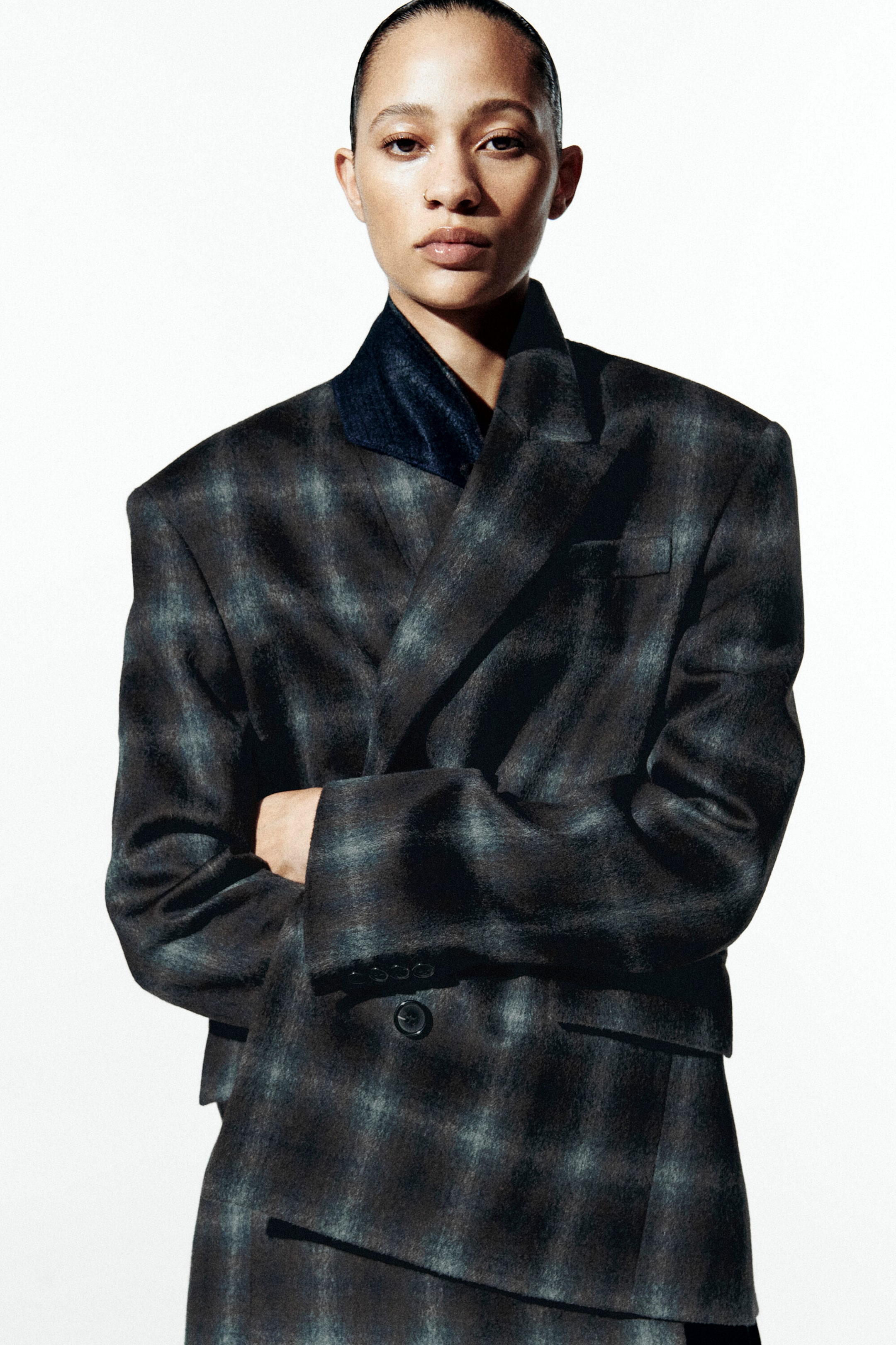 Un blazer à coupe décontractée, croisé, dans un motif à carreaux gris foncé et bleu diffus, est présenté de face, avec des revers en pointe et des épaules structurées. Le blazer est porté par-dessus une chemise sombre à col, mettant en valeur sa silhouette oversize et sa fermeture à boutons.