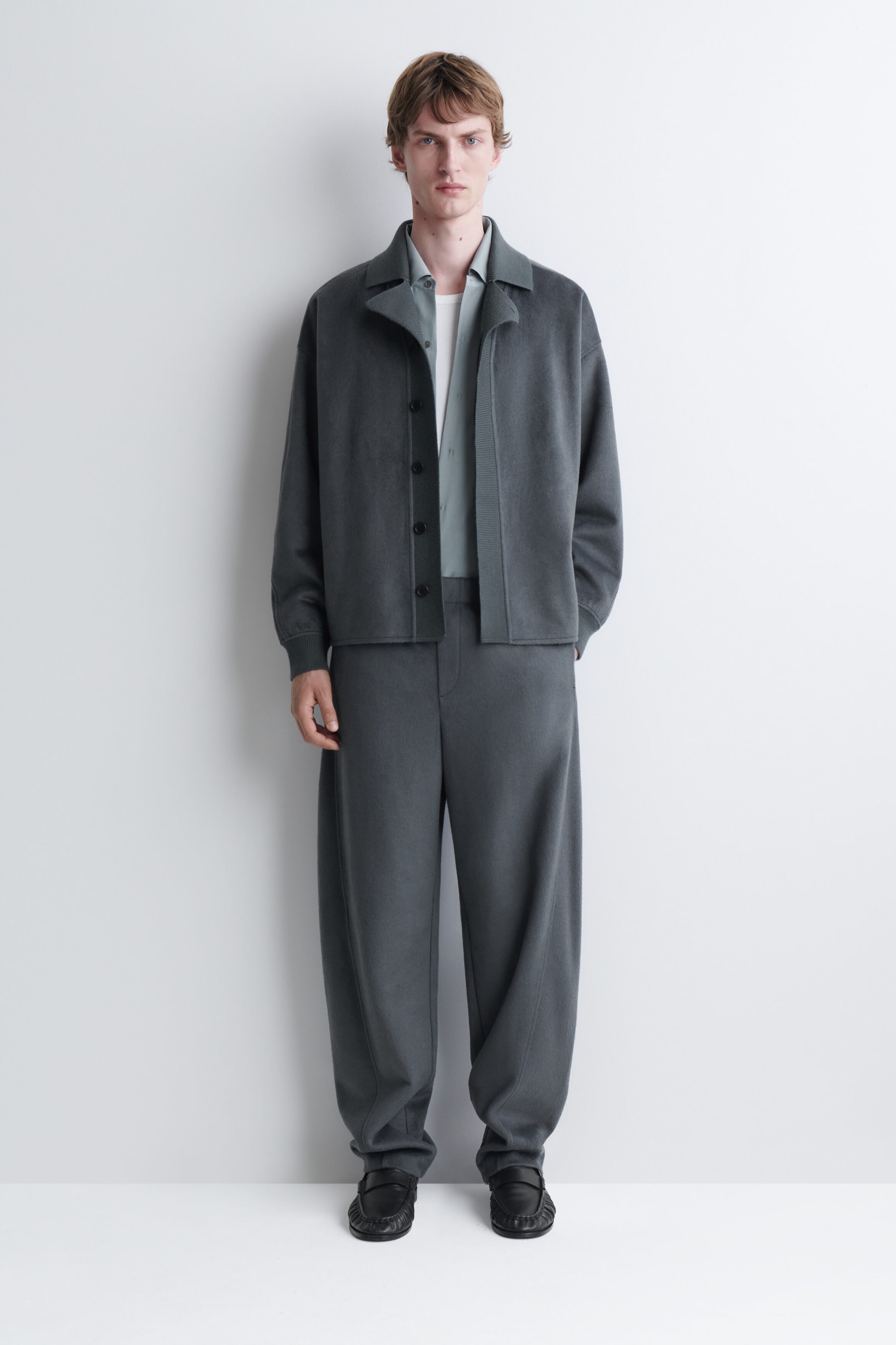 Le mannequin porte une veste en tricot gris foncé et un pantalon ample assorti. La veste ouverte présente un large col et est superposée sur une chemise gris pâle à col et un t-shirt blanc.