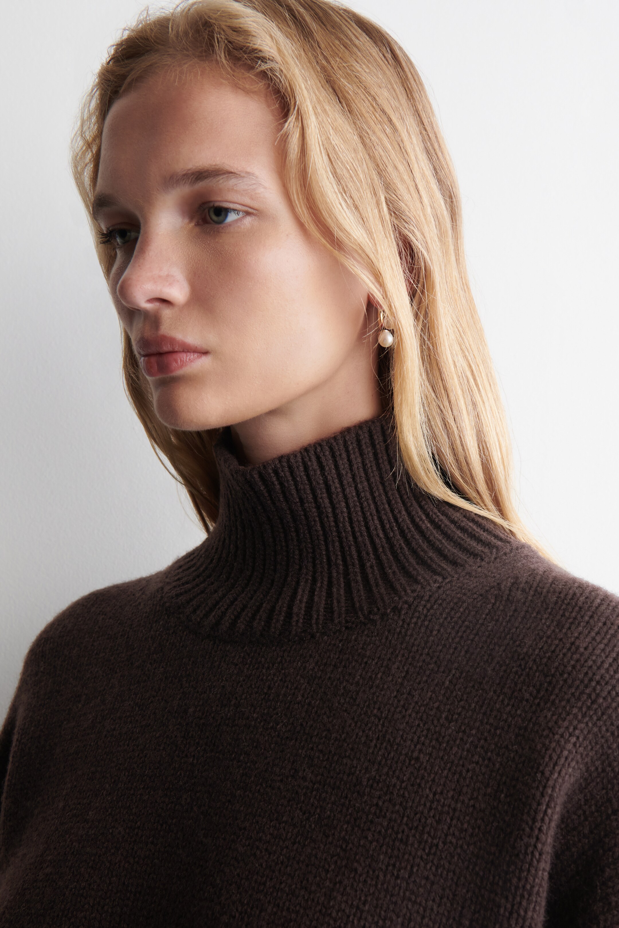 View larger image: A dark brown turtleneck sweater is featuring a high, ribbed collar. The sweater is showcasing a fine knit texture on its body and is presented from a slight side-front angle, highlighting the collar's structure and fit around the neck.