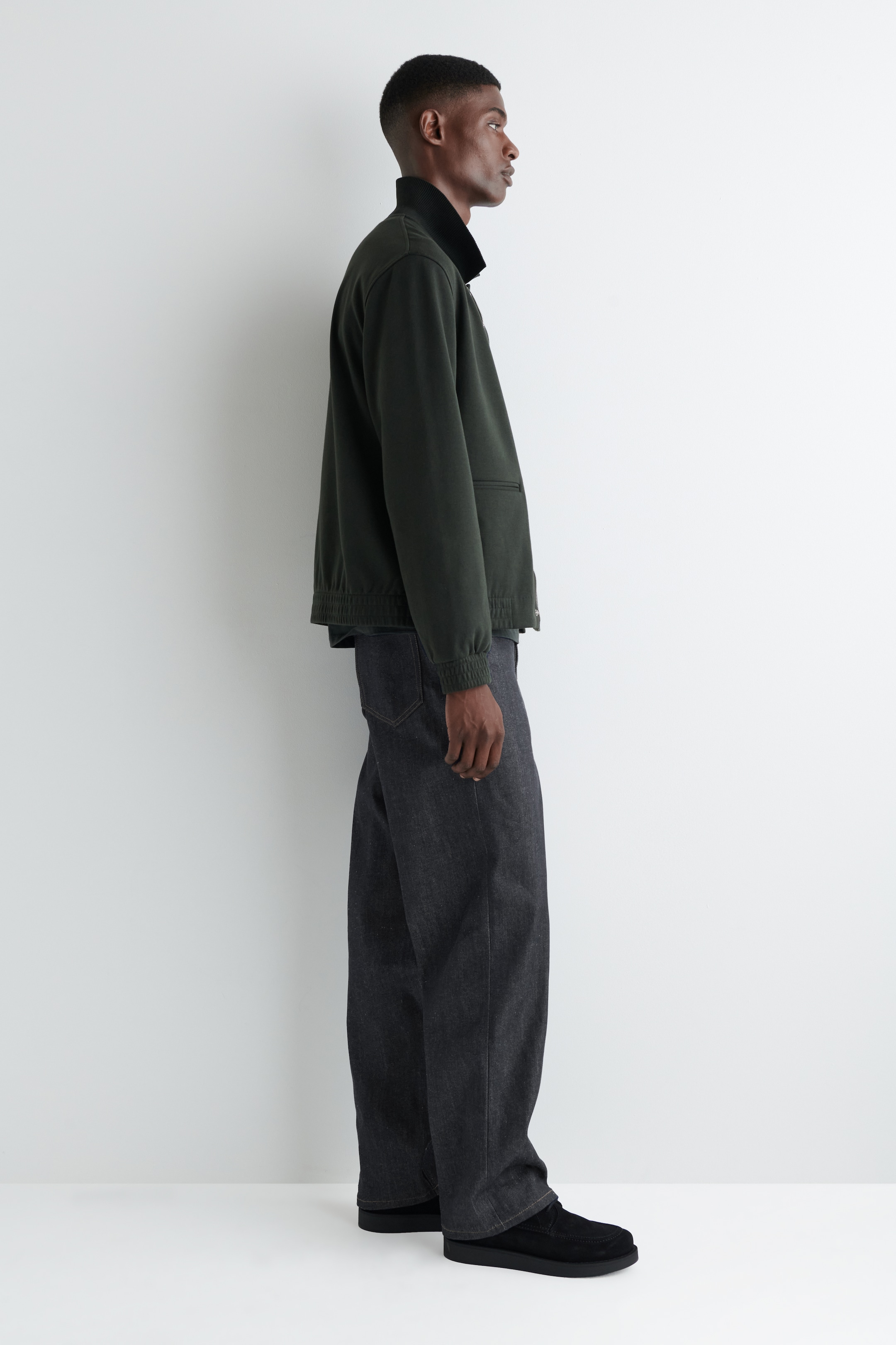 Visualizza immagine più grande: Una giacca casual verde scuro viene presentata di profilo, mettendo in risalto il colletto nero rialzato, la chiusura frontale con zip completa e l'orlo e i polsini a coste, abbinata a jeans a gamba larga con lavaggio scuro.