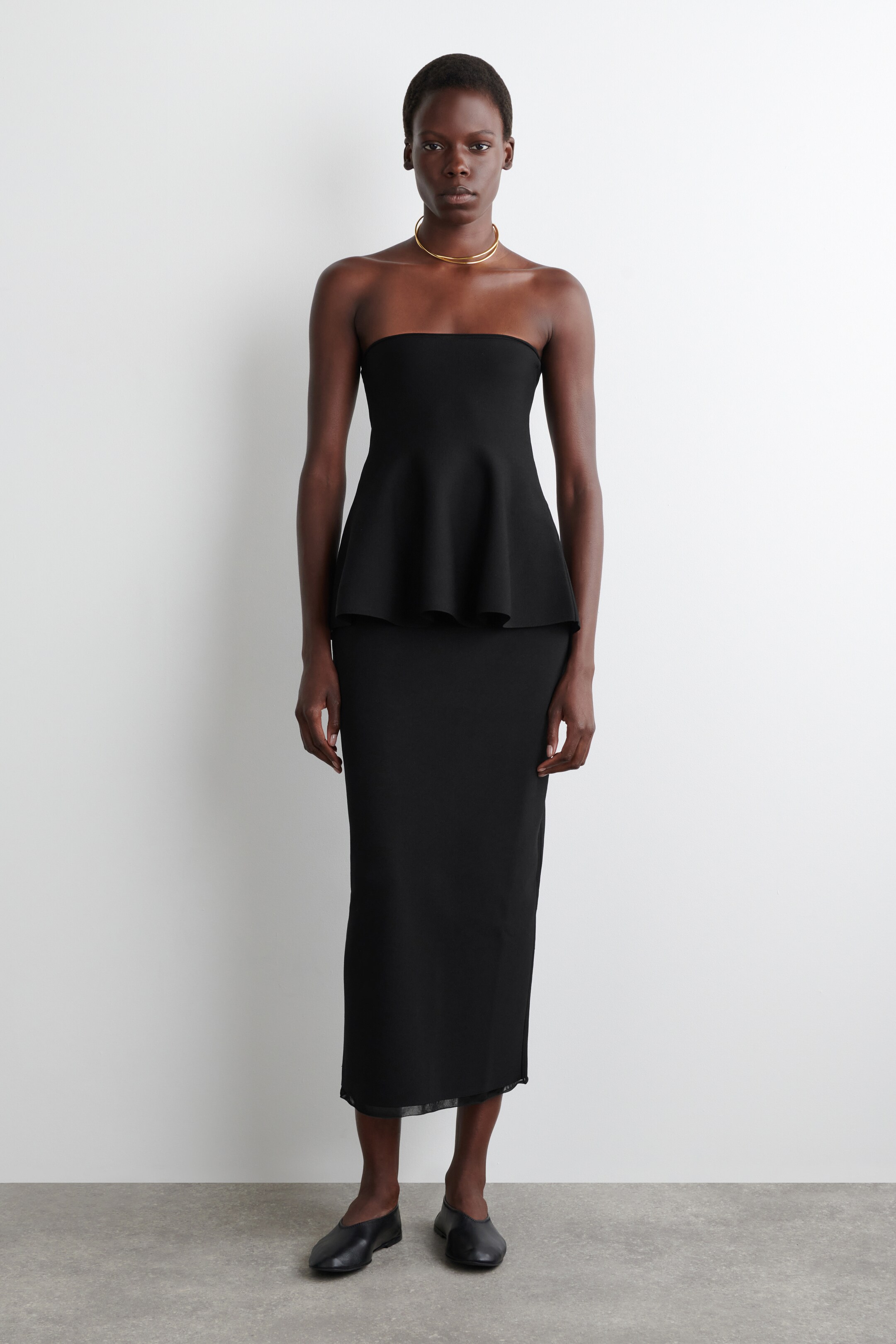 Ver imagen más grande: Un vestido negro sin tirantes de corte peplum presenta una silueta ajustada, mirando hacia adelante, con un corpiño estructurado y un peplum que se ensancha desde la cintura. La falda recta de longitud midi tiene un ribete transparente en su dobladillo.