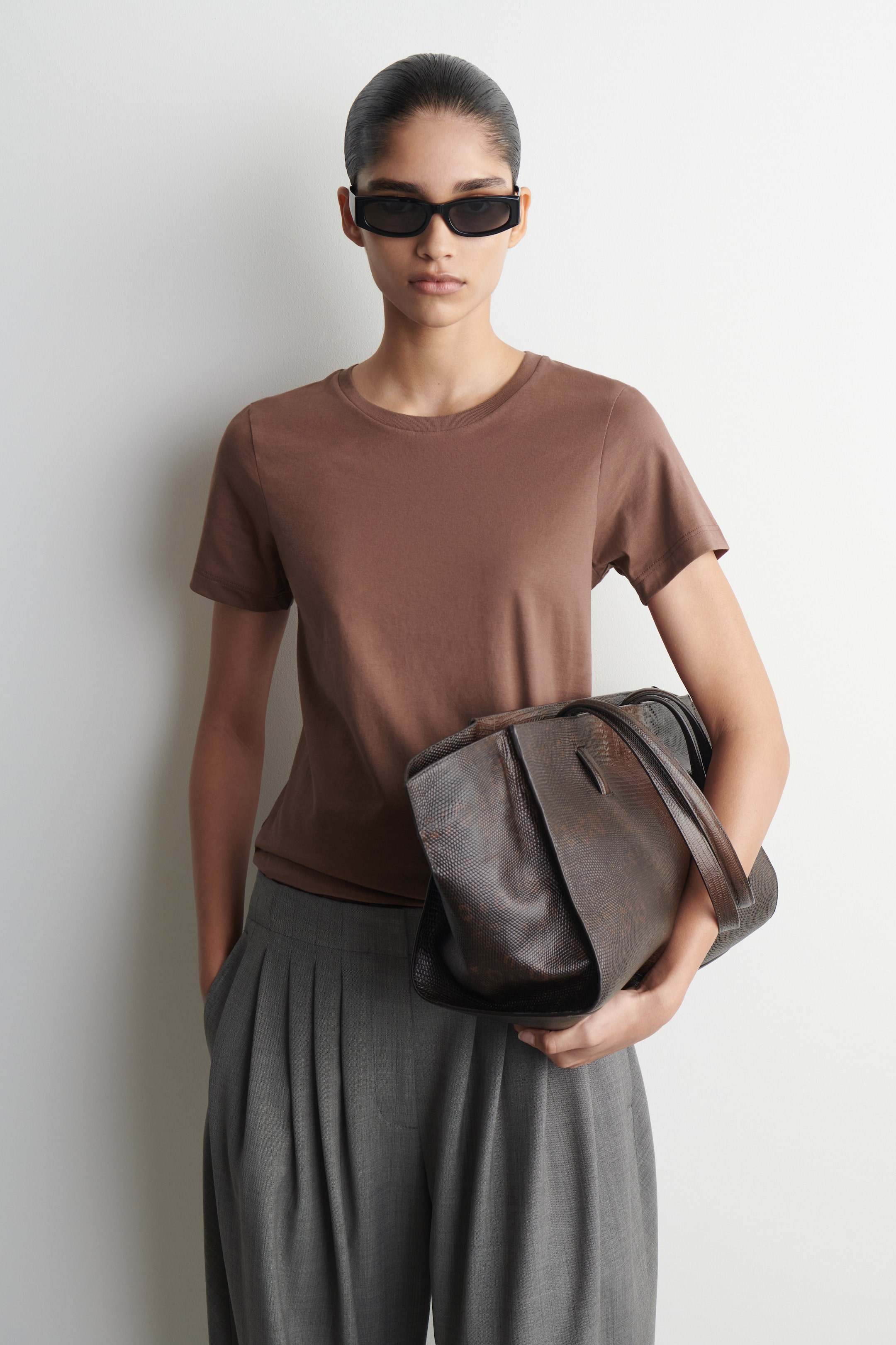 Zobraziť väčší obrázok: Hnedé tričko s krátkym rukávom je nosené, prezentované z mierneho predného pravého uhla, pričom ukazuje svoj okrúhly výstrih a hladkú, jednoduchú textúru. Má voľný strih, jemne sa splýva cez trup.