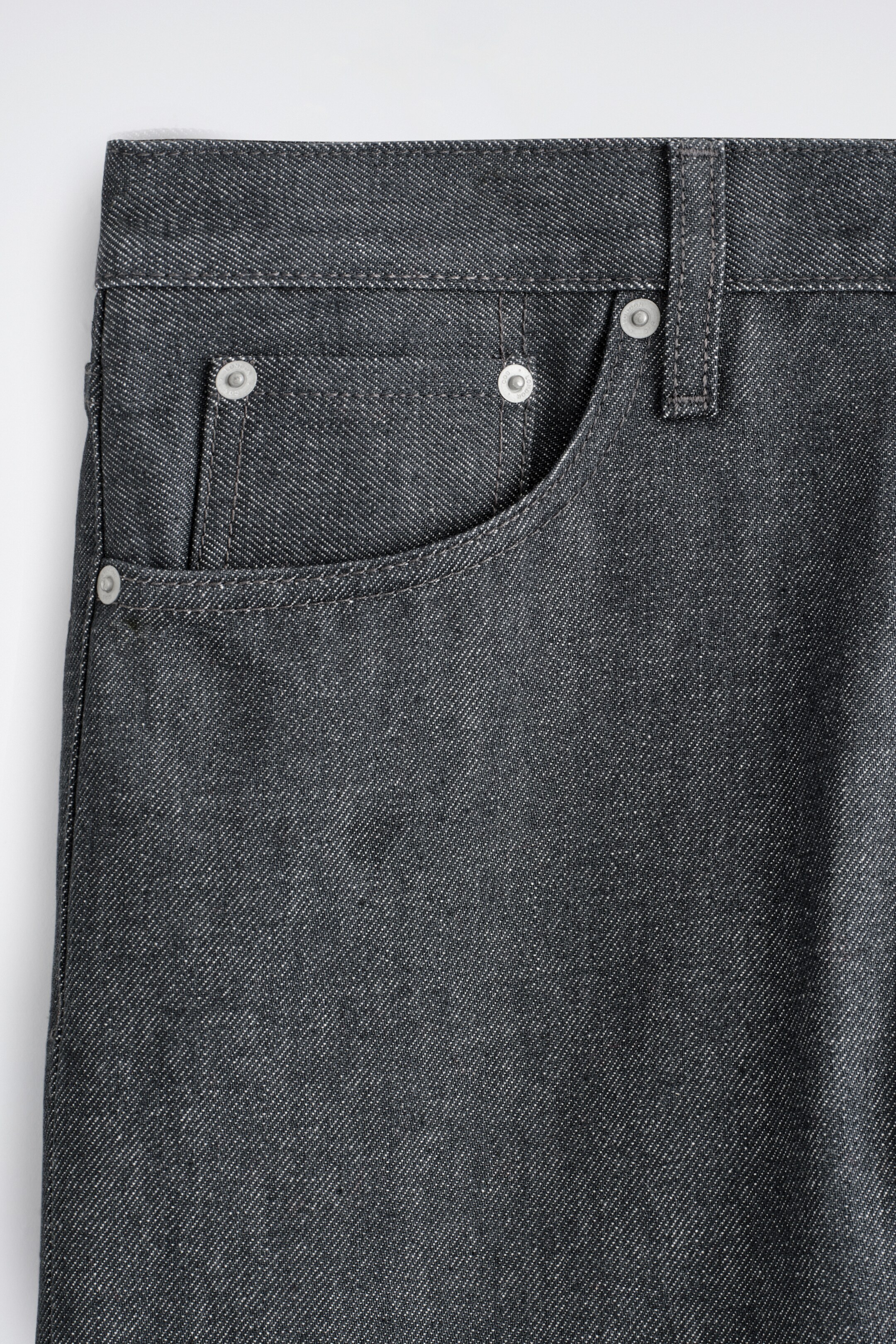 Agrandir l'image: L'AVANT du jean en denim gris foncé présente une texture sergée visible, une poche avant incurvée et une petite poche à monnaie, toutes renforcées par des rivets de couleur argent et détaillées avec des surpiqûres contrastantes le long des poches et de la ceinture.