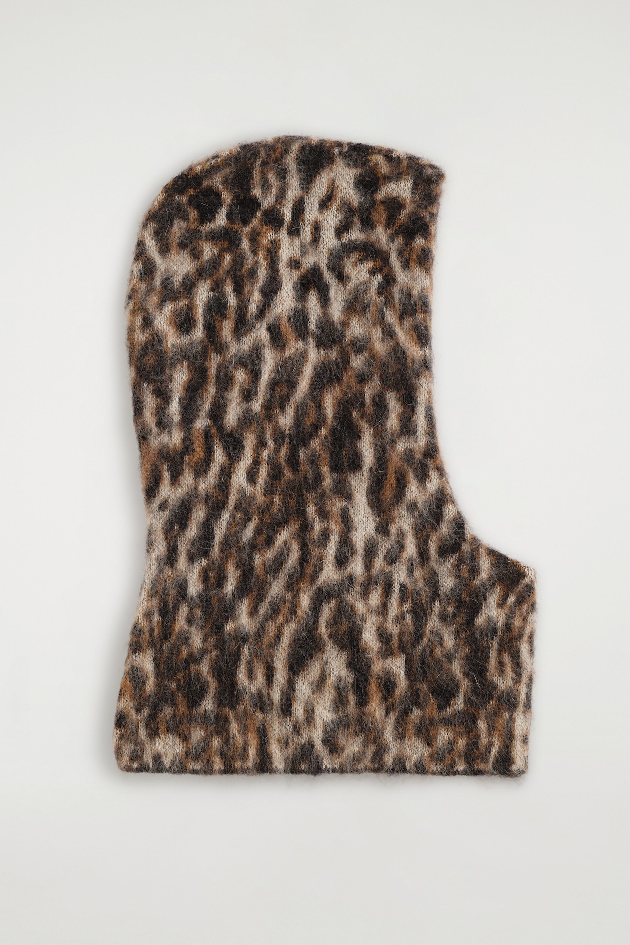 A knit balaclava featuring a brown and black leopard print is lying flat in profile facing right, showcasing a fuzzy, brushed texture. The hood displays a curved head shape, a straight bottom hem, and a cut-out opening for the face on the right side.
