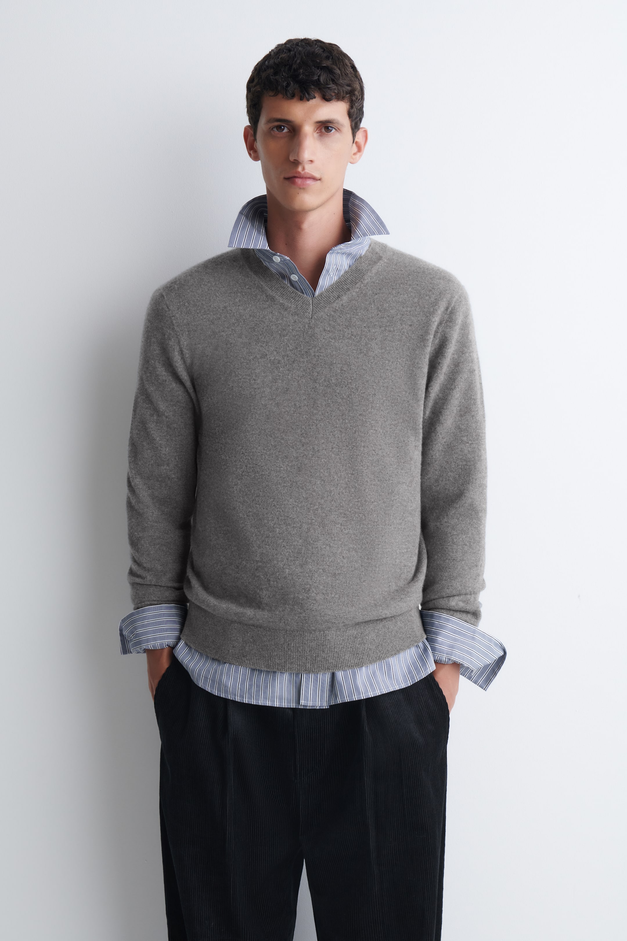 Visualizza l'immagine ingrandita: Il modello indossa un maglione grigio in cashmere con scollo a V, collo, polsini e bordo a coste. Il maglione è abbinato a una camicia con colletto a righe blu e bianche ed è indossato sopra un paio di pantaloni neri.