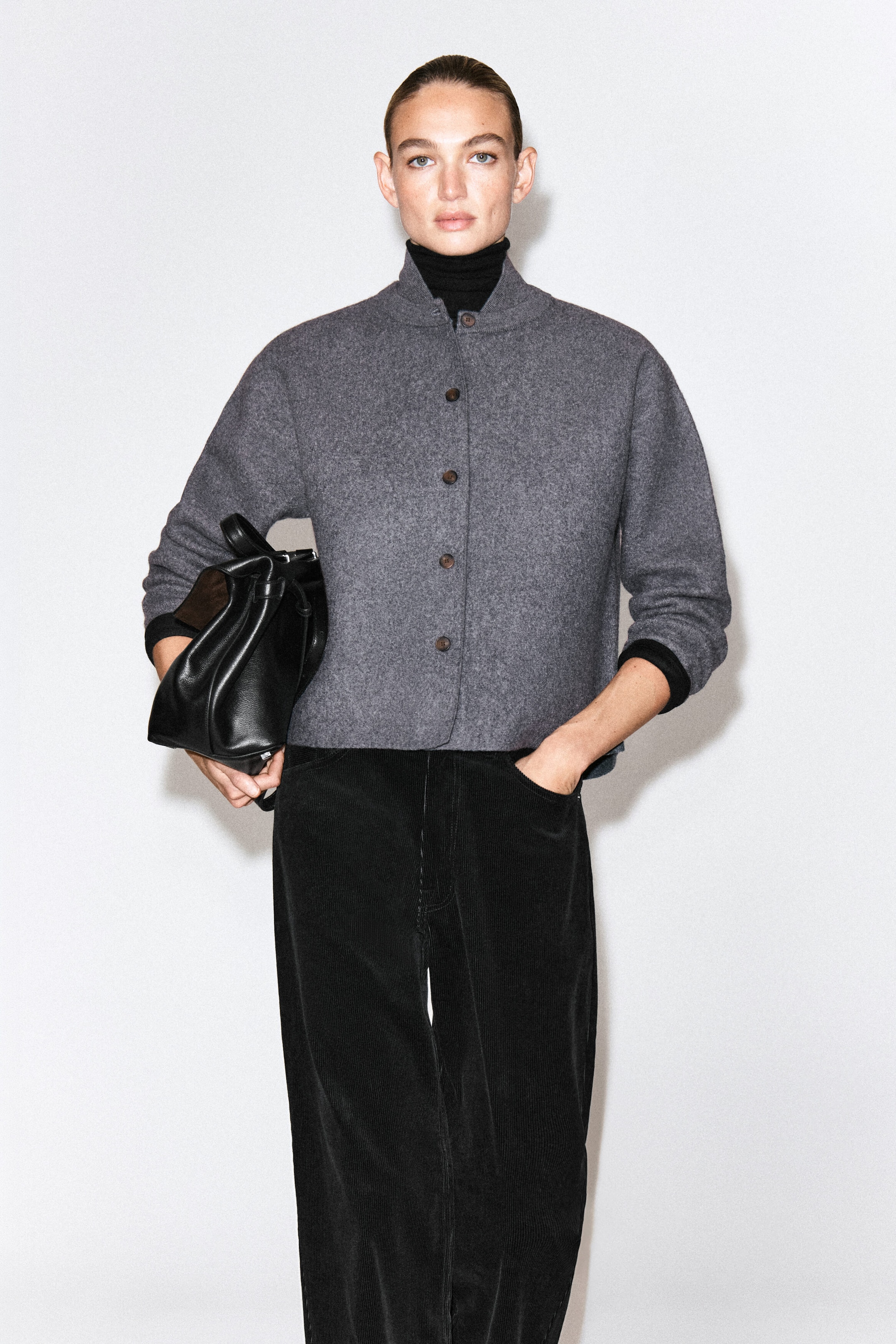 A gray boiled wool cardigan with a rounded silhouette and button-front closure is worn by the model over a black turtleneck. The cardigan is styled with black corduroy pants, and a black leather bag is carried under the arm. A gray boiled wool cardigan with a rounded silhouette and button-front closure is worn by the model over a black turtleneck. The cardigan is styled with black corduroy pants, and a black leather bag is carried under the arm.