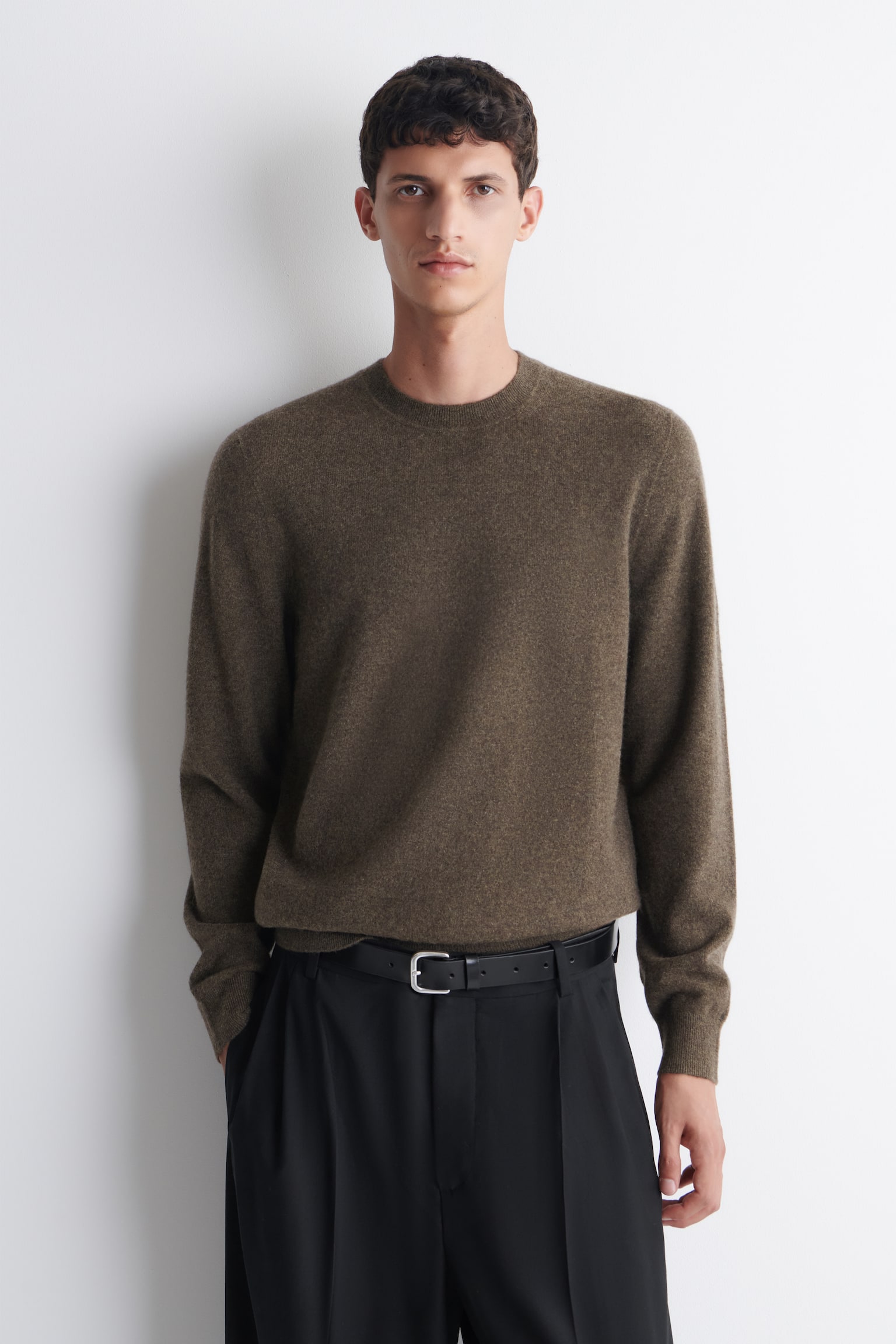 Un pull à col rond marron moyen est posé à plat, face avant visible, présentant une texture en maille fine, des manches longues avec poignets côtelés et un ourlet côtelé. Une étiquette blanche affichant « COS » est visible à l’arrière du col. - 1
