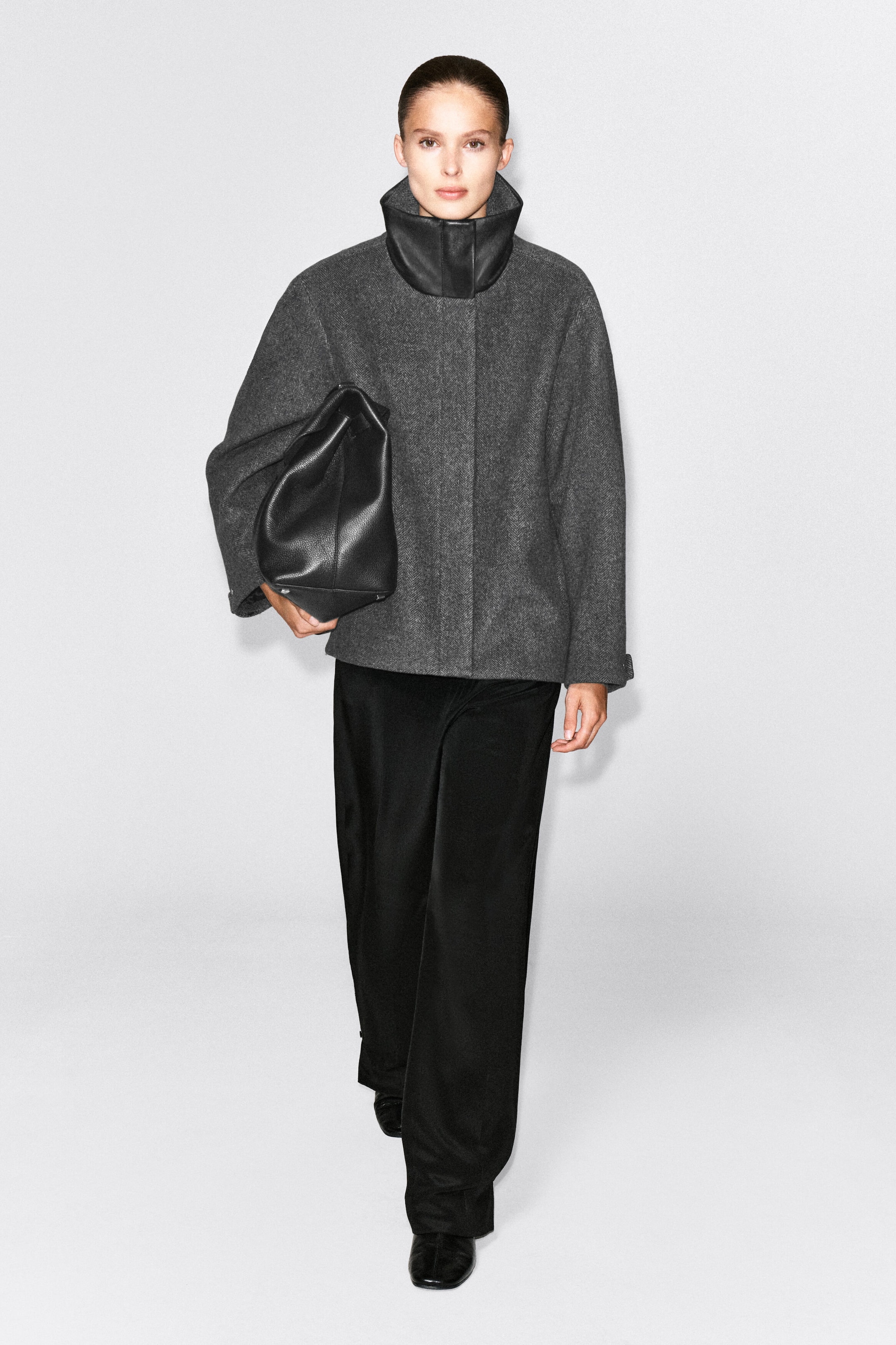 Ein oversized grauer Jacke aus Wollmischung mit schwarzem Leder-Stehkragen wird vom Model getragen. Die Jacke ist mit langen schwarzen Hosen kombiniert, und eine grosse schwarze Ledertasche wird unter dem Arm getragen. Ein oversized grauer Jacke aus Wollmischung mit schwarzem Leder-Stehkragen wird vom Model getragen. Die Jacke ist mit langen schwarzen Hosen kombiniert, und eine grosse schwarze Ledertasche wird unter dem Arm getragen.