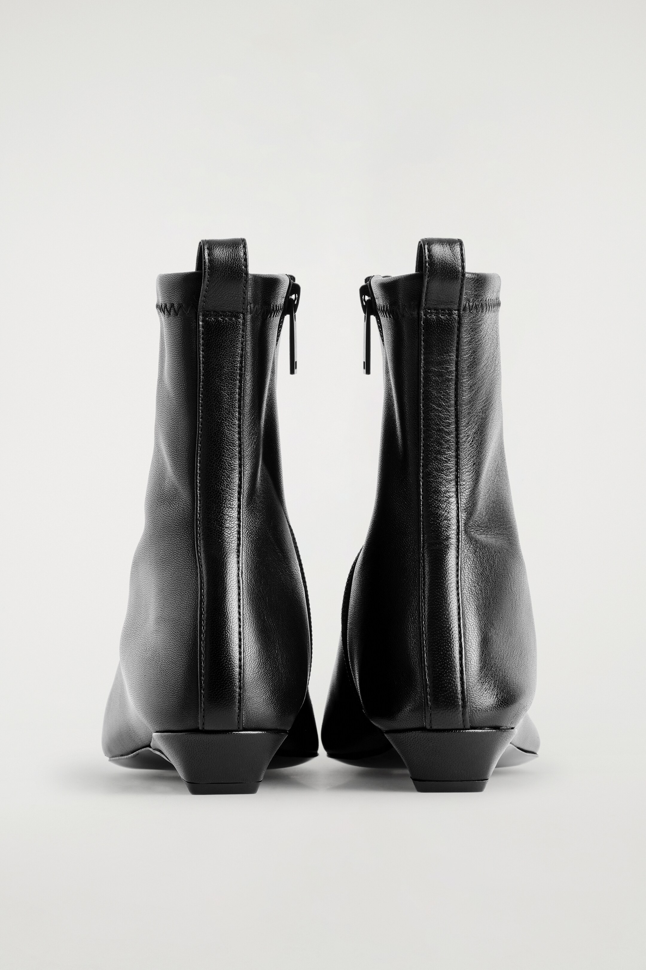 Größeres Bild ansehen: Spitze schwarze Leder-Sockenstiefel werden von hinten betrachtet, mit glattem Leder im unteren Teil und einem dehnbaren, gerippten oberen Schaft. Ein Reißverschluss verläuft vertikal den Rücken jedes Stiefels hinauf, gekrönt mit einem Zuglasche, und jeder Stiefel ruht auf einem niedrigen, konischen Absatz.