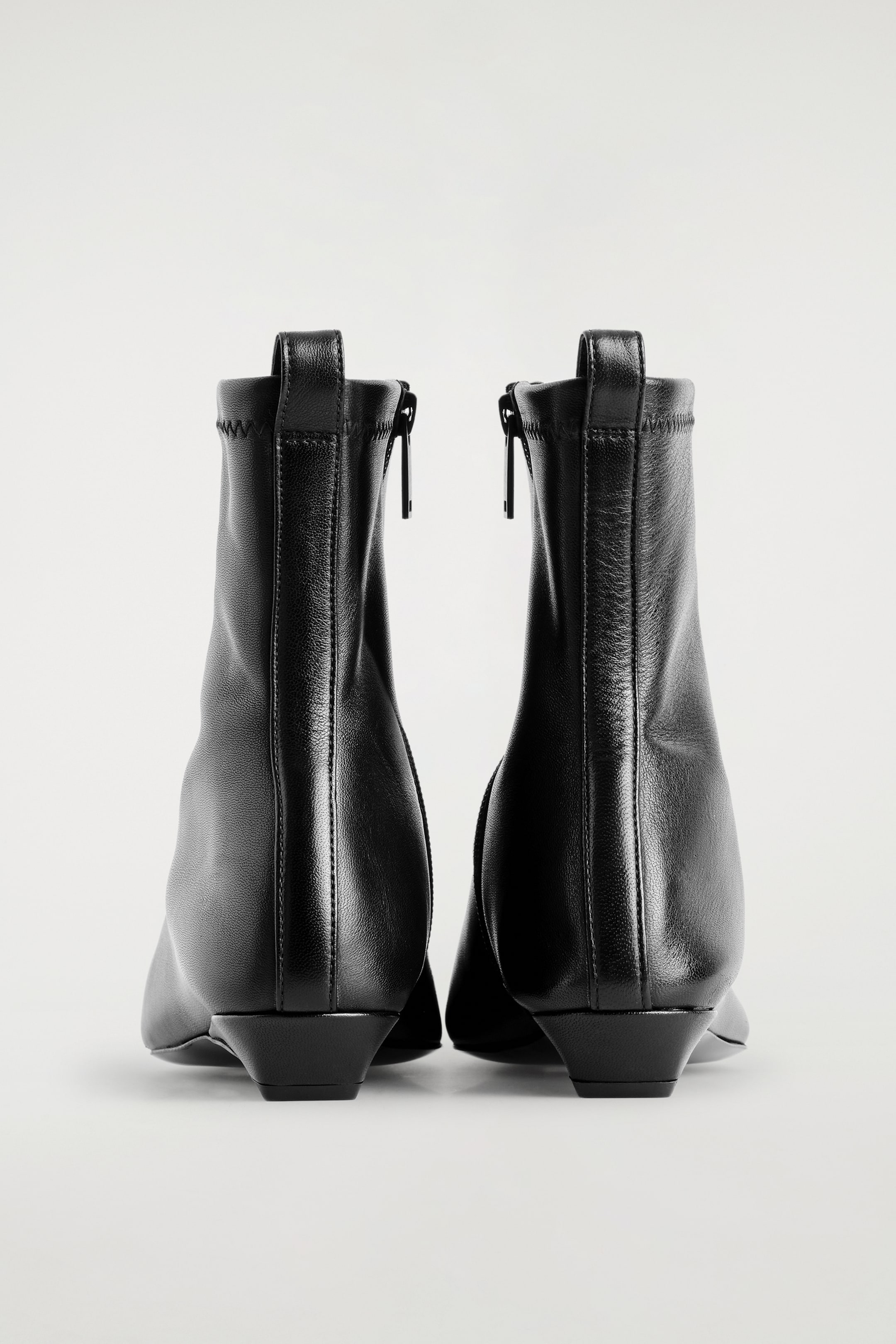 Agrandir l'image: Des bottines chaussettes en cuir noir pointu sont vues de l'arrière, avec du cuir lisse sur la partie inférieure et une tige supérieure extensible et côtelée. Une fermeture éclair monte verticalement à l'arrière de chaque botte, surmontée d'une languette, et chaque botte repose sur un talon bas et effilé.