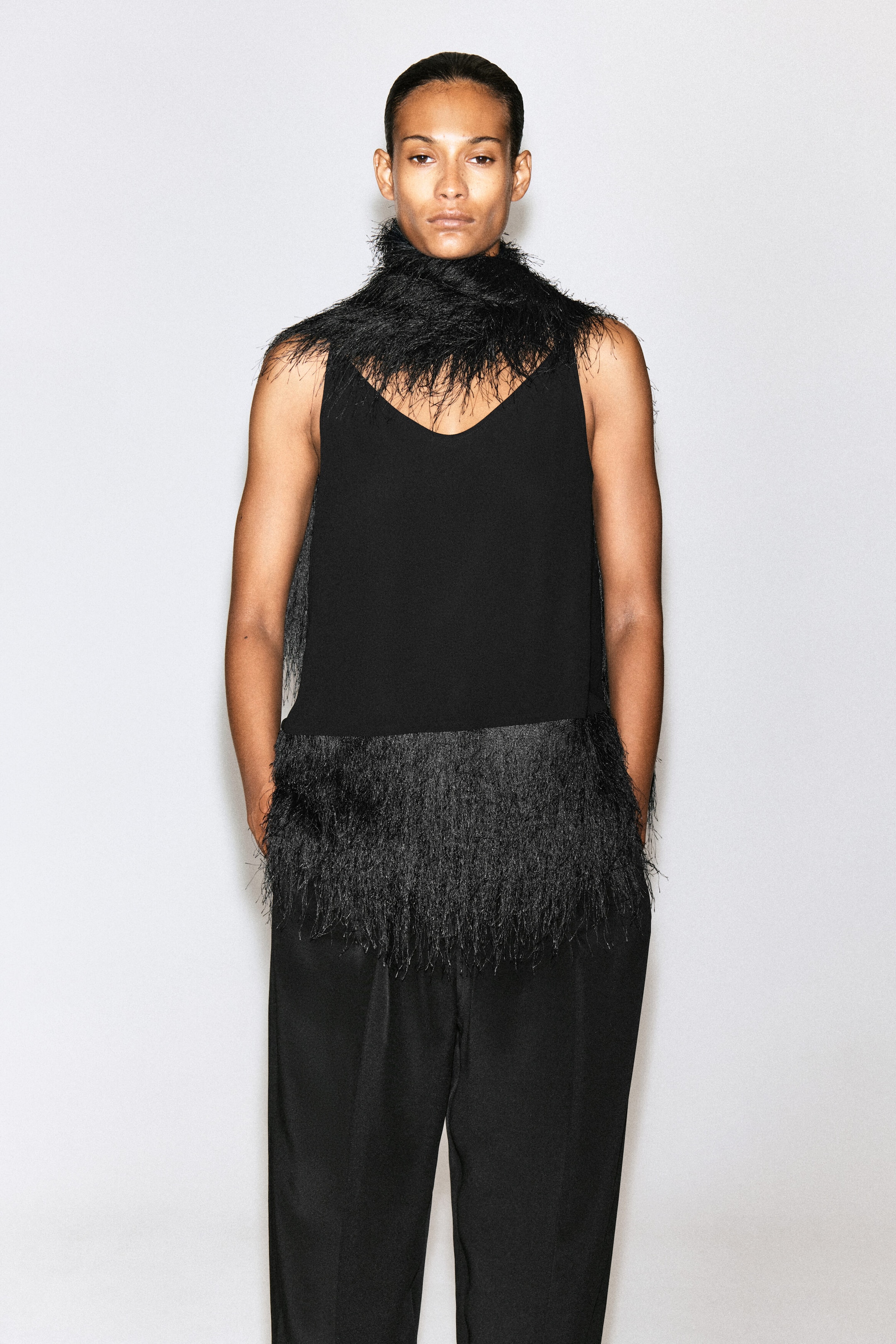 A black scarf with wispy, feather-like fringe is worn wrapped closely around the model’s neck. The accessory is styled over a black sleeveless top and black trousers.