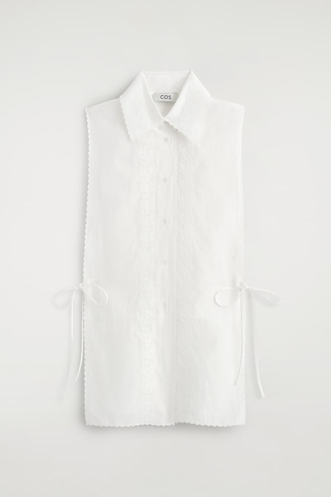 Un plastron blanc brodé à bord festonné est présenté de face, avec un col pointu à bords festonnés et une patte de boutonnage sur le devant. Il présente des panneaux de dentelle brodée verticaux de part et dâautre des boutons, une bordure festonnée sur tous les bords extérieurs, ainsi que de fins liens à nouer sur les côtés. - 1