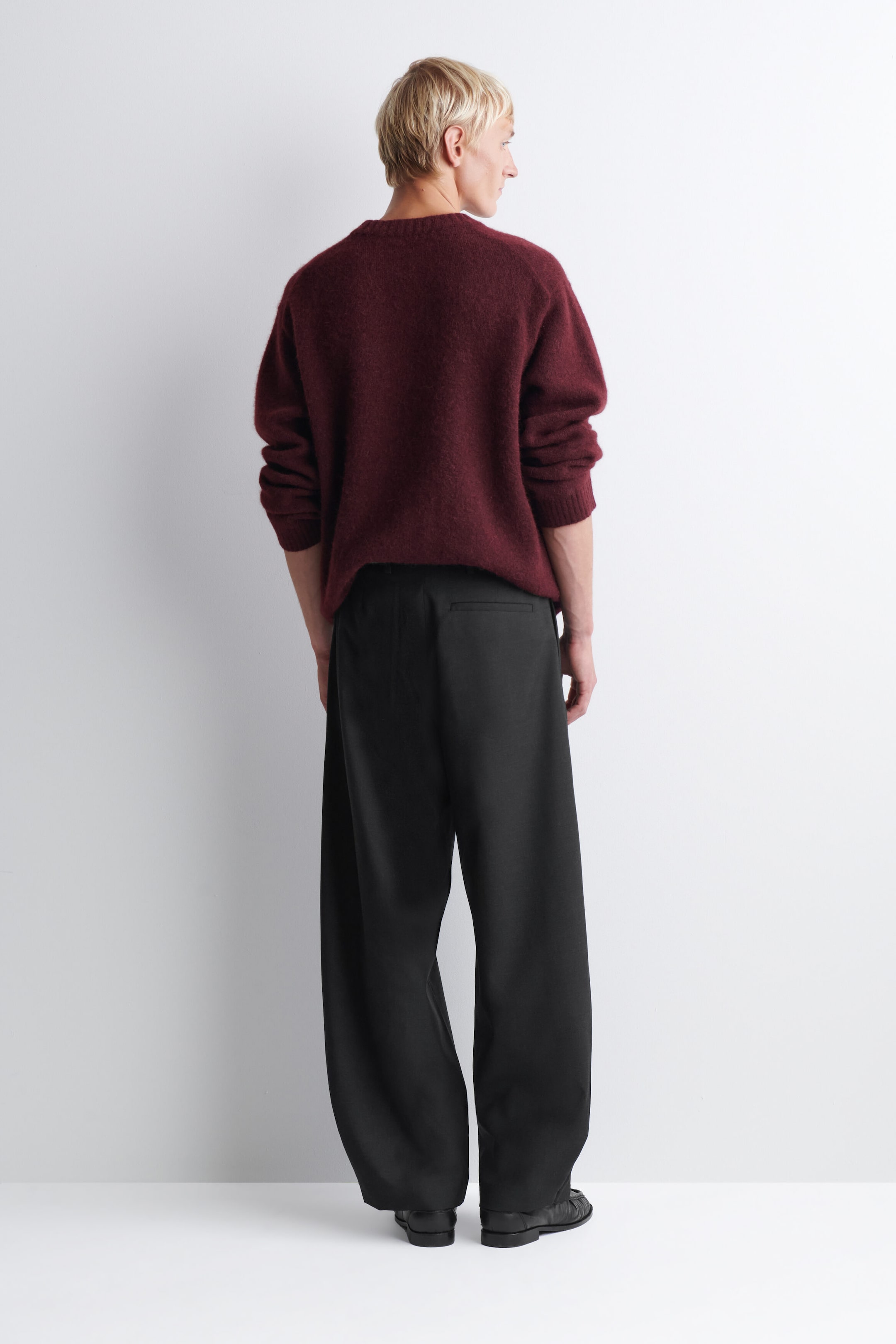 The belt-detail wool relaxed-fit barrel-leg trousers are shown from the back, highlighting their loose, curved silhouette in black wool fabric. The trousers drape loosely down the legs, featuring a single welt pocket on the right side and pooling slightly over black leather shoes, while a burgundy knit jumper is draped over the waistband. The belt-detail wool relaxed-fit barrel-leg trousers are shown from the back, highlighting their loose, curved silhouette in black wool fabric. The trousers drape loosely down the legs, featuring a single welt pocket on the right side and pooling slightly over black leather shoes, while a burgundy knit jumper is draped over the waistband.
