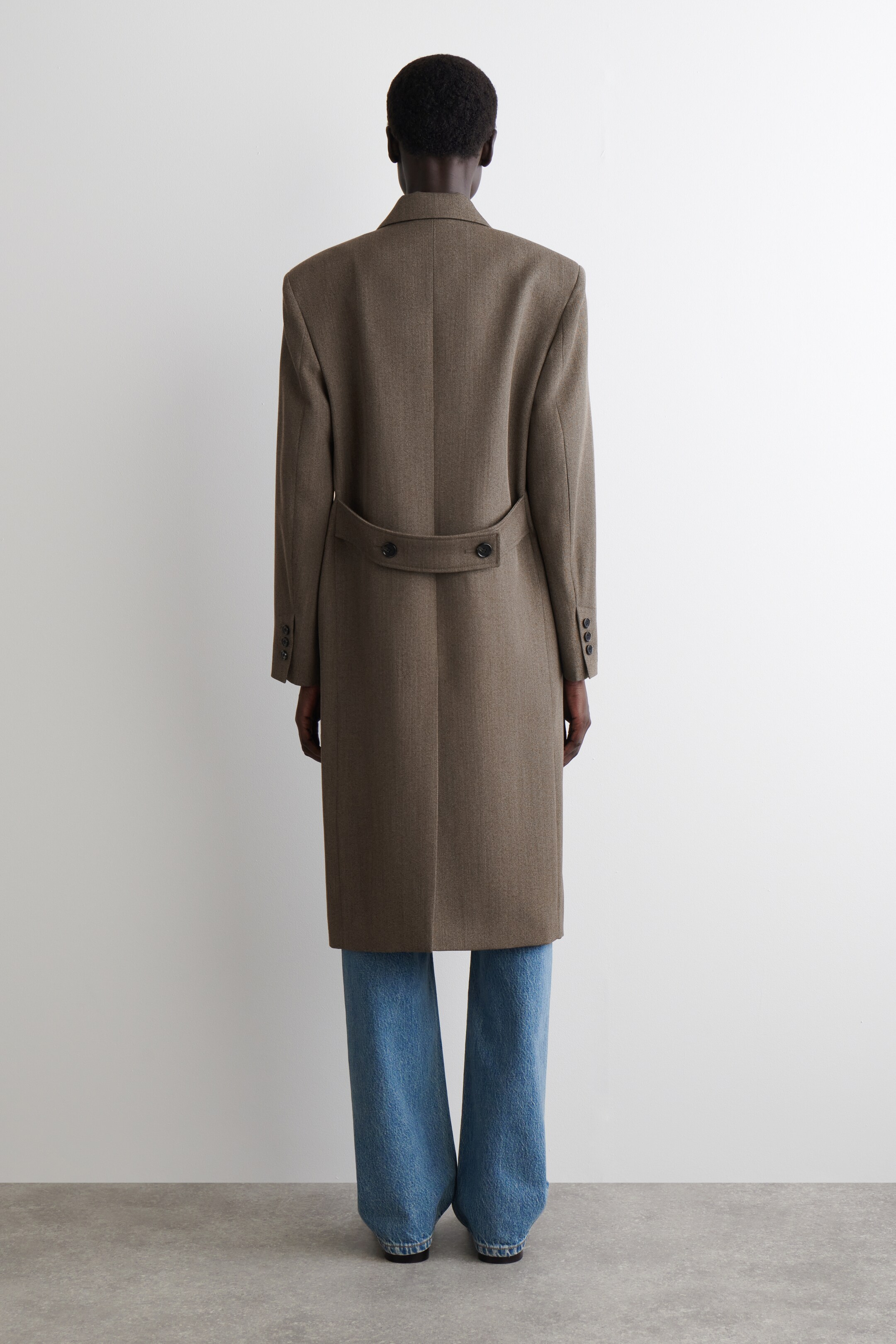 Agrandir l'image: Un long manteau marron à chevrons avec des épaules structurées est présenté de dos, s'étendant sous les genoux. Il présente une demi-ceinture boutonnée à l'arrière de la taille, une seule fente arrière, et trois boutons sur chaque manchette, et est porté par-dessus un jean bleu clair.