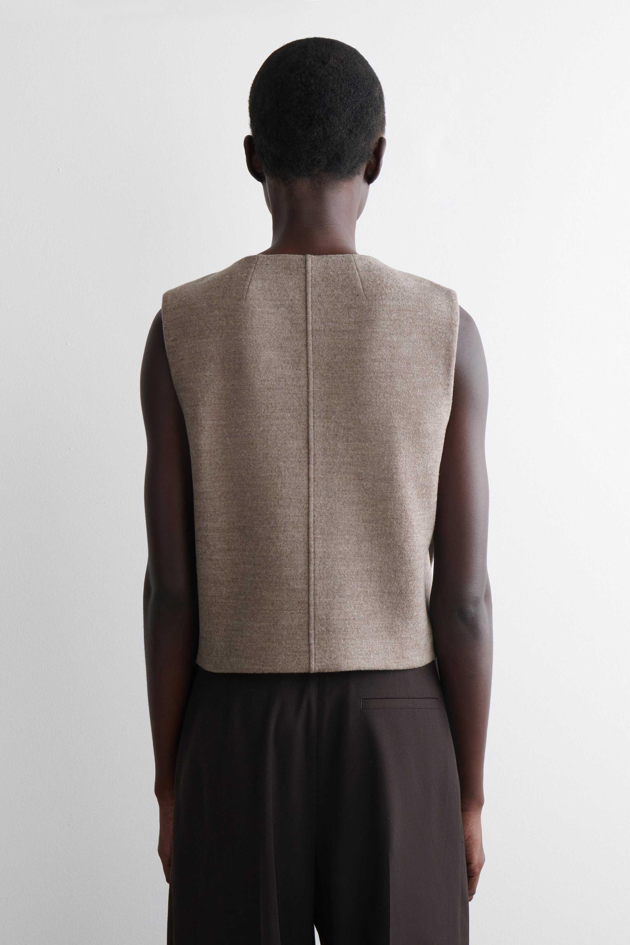 Agrandir l'image: Un gilet sans manches brun clair est vu de dos, avec une couture centrale verticale proéminente, des coutures d'épaule angulaires et un tissu texturé avec une longueur courte, se terminant à la taille sur un pantalon brun foncé.