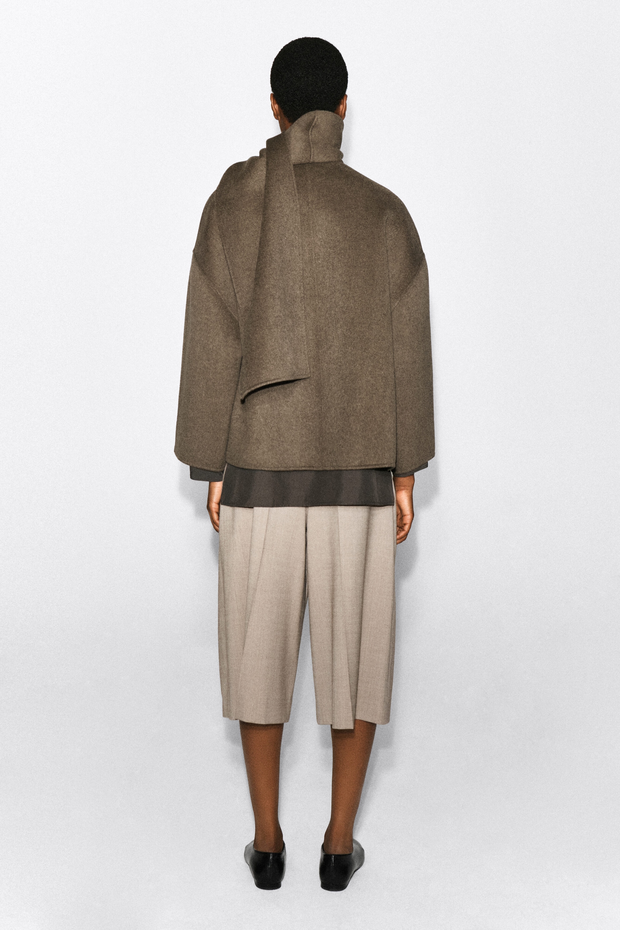 A double-faced wool-blend scarf jacket in a muted brown is being worn by the model, shown from the back. The jacket features a relaxed fit with an integrated scarf draped over the shoulder, layered over a dark grey top and light beige shorts. A double-faced wool-blend scarf jacket in a muted brown is being worn by the model, shown from the back. The jacket features a relaxed fit with an integrated scarf draped over the shoulder, layered over a dark grey top and light beige shorts.