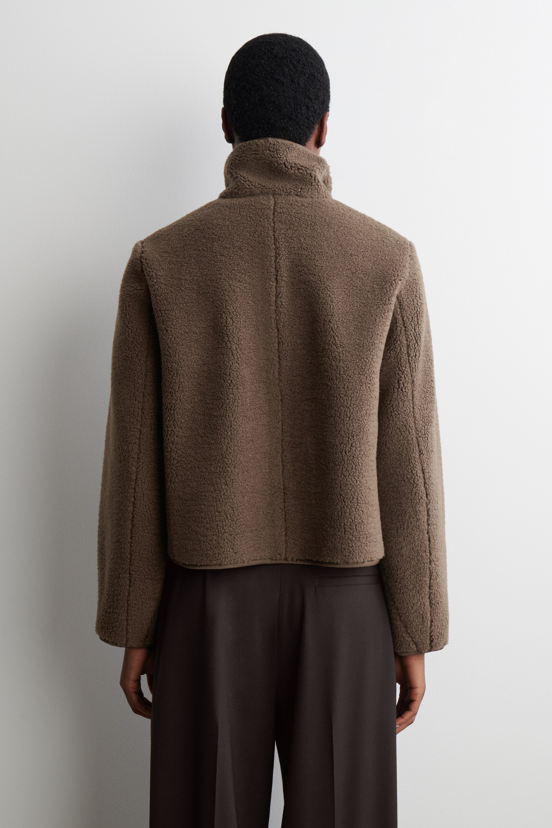 Agrandir l'image: Une veste en peluche de nounours marron est présentée de dos, avec un haut col montant. La veste est courte, et une couture verticale descend le centre du dos.