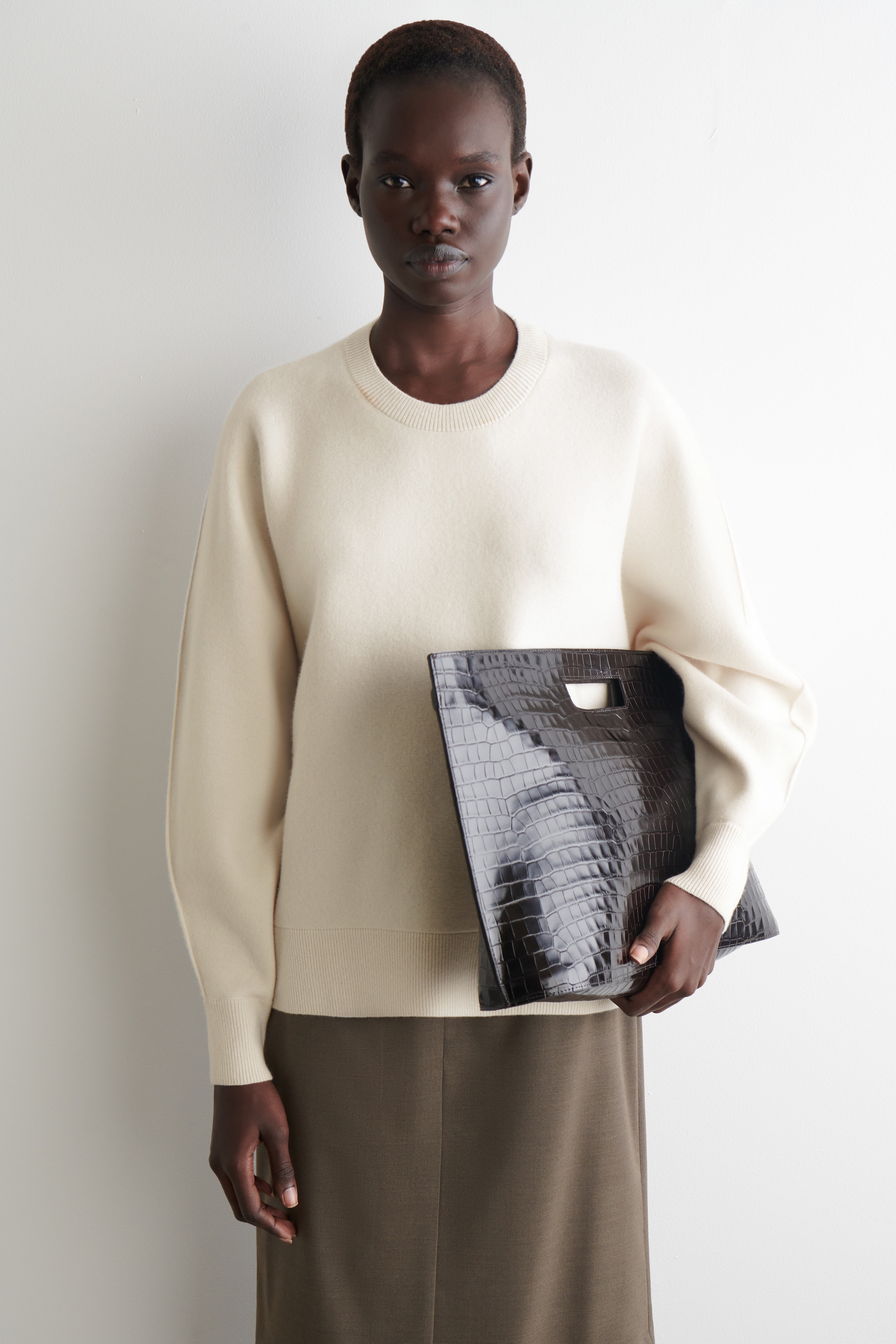 Visualizza immagine più grande: Un maglione girocollo color bianco sporco è indossato, caratterizzato da una vestibilità rilassata con spalle scese e polsini e orlo a coste. Una pochette marrone scuro con stampa coccodrillo e manico intagliato è tenuta in mano, apparendo davanti al maglione, che è abbinato a una gonna midi dritta color oliva marrone.