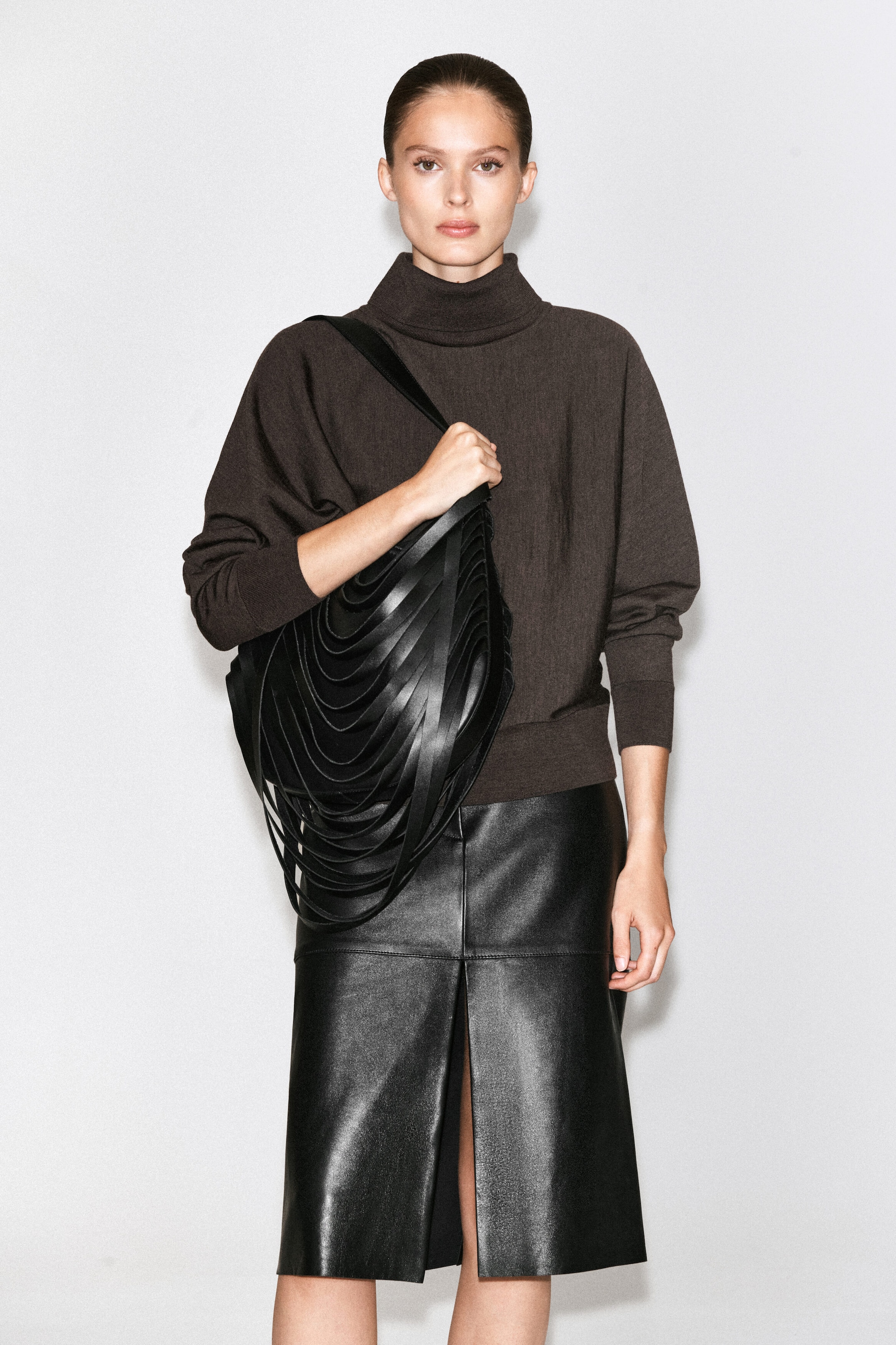A black leather shoulder bag featuring a sculptural design with cascading, draped loops is being carried by the model. The model is holding the wide strap of the bag, styled over a dark brown roll-neck jumper and paired with a black leather midi skirt. A black leather shoulder bag featuring a sculptural design with cascading, draped loops is being carried by the model. The model is holding the wide strap of the bag, styled over a dark brown roll-neck jumper and paired with a black leather midi skirt.