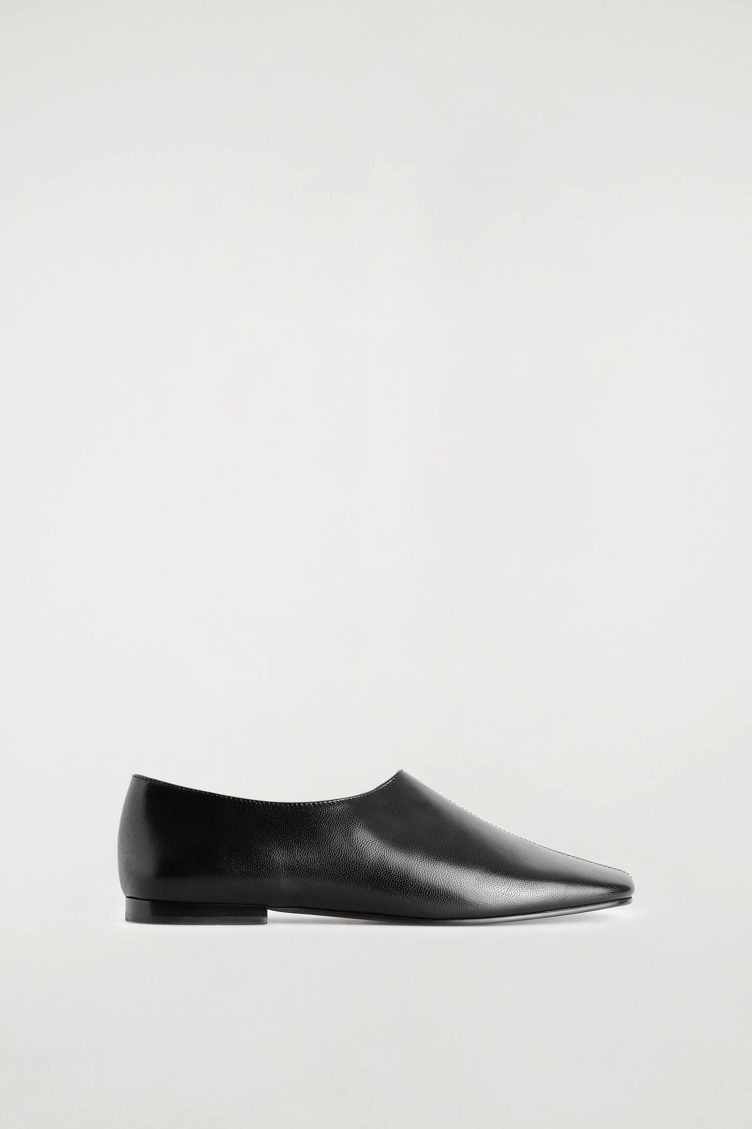 Un Ãºnico zapato plano de cuero negro aparece apoyado en vista de perfil lateral, orientado hacia la derecha, mostrando su puntera cuadrada con una discreta costura central a pespunte en la parte superior. El zapato presenta un escote alto y un tacÃ³n bajo y plano.