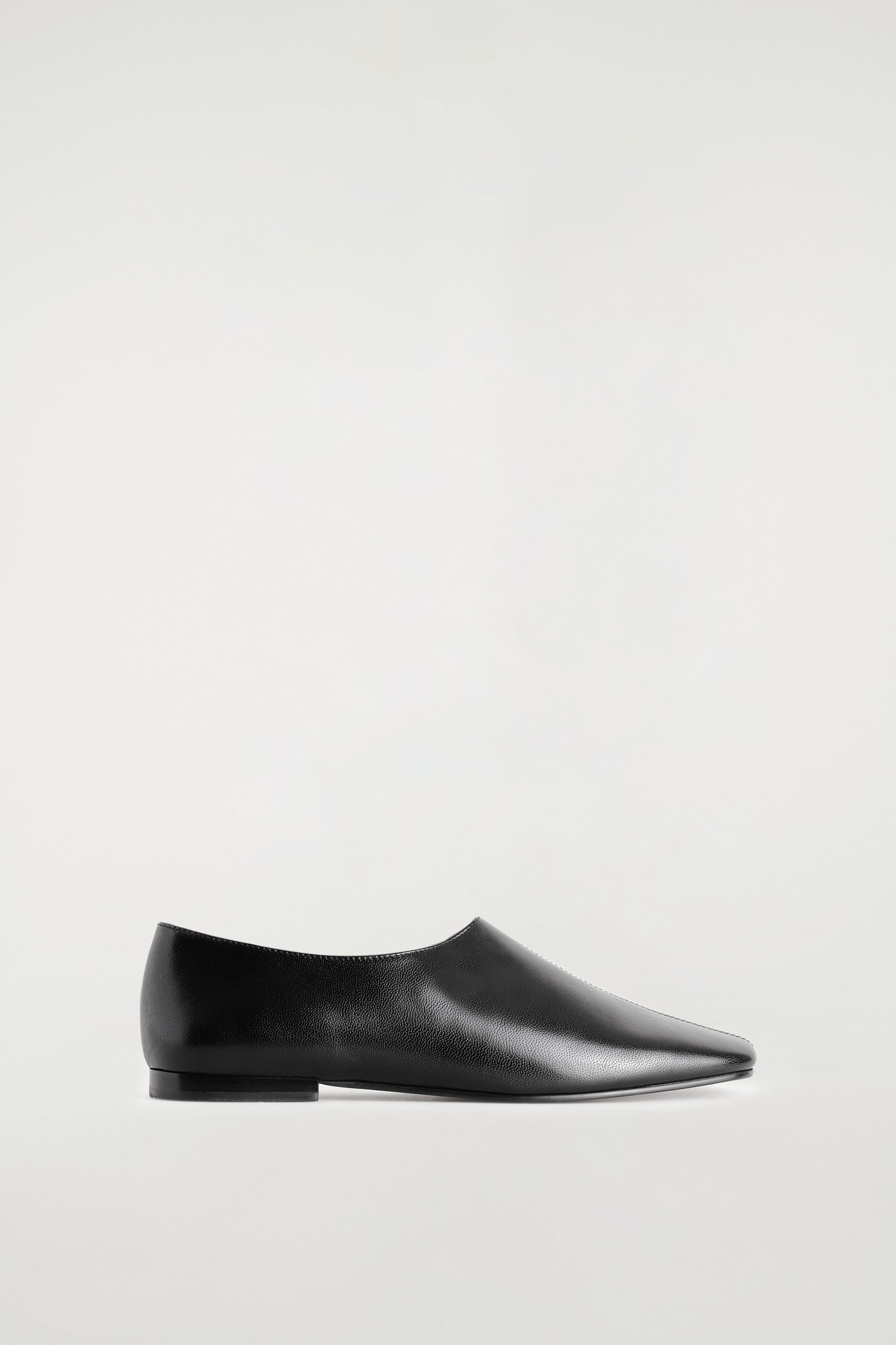 Ver imagen más grande: Un único zapato plano de cuero negro aparece apoyado en vista de perfil lateral, orientado hacia la derecha, mostrando su puntera cuadrada con una discreta costura central a pespunte en la parte superior. El zapato presenta un escote alto y un tacón bajo y plano.