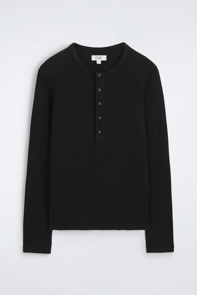 Un t-shirt henley noir, ajusté et côtelé en coton à manches longues est présenté à plat, montrant son design de face. Le vêtement présente un col rond avec une patte de boutonnage verticale à cinq boutons ainsi quâune fine texture côtelée sur lâensemble du corps et des manches. - 1