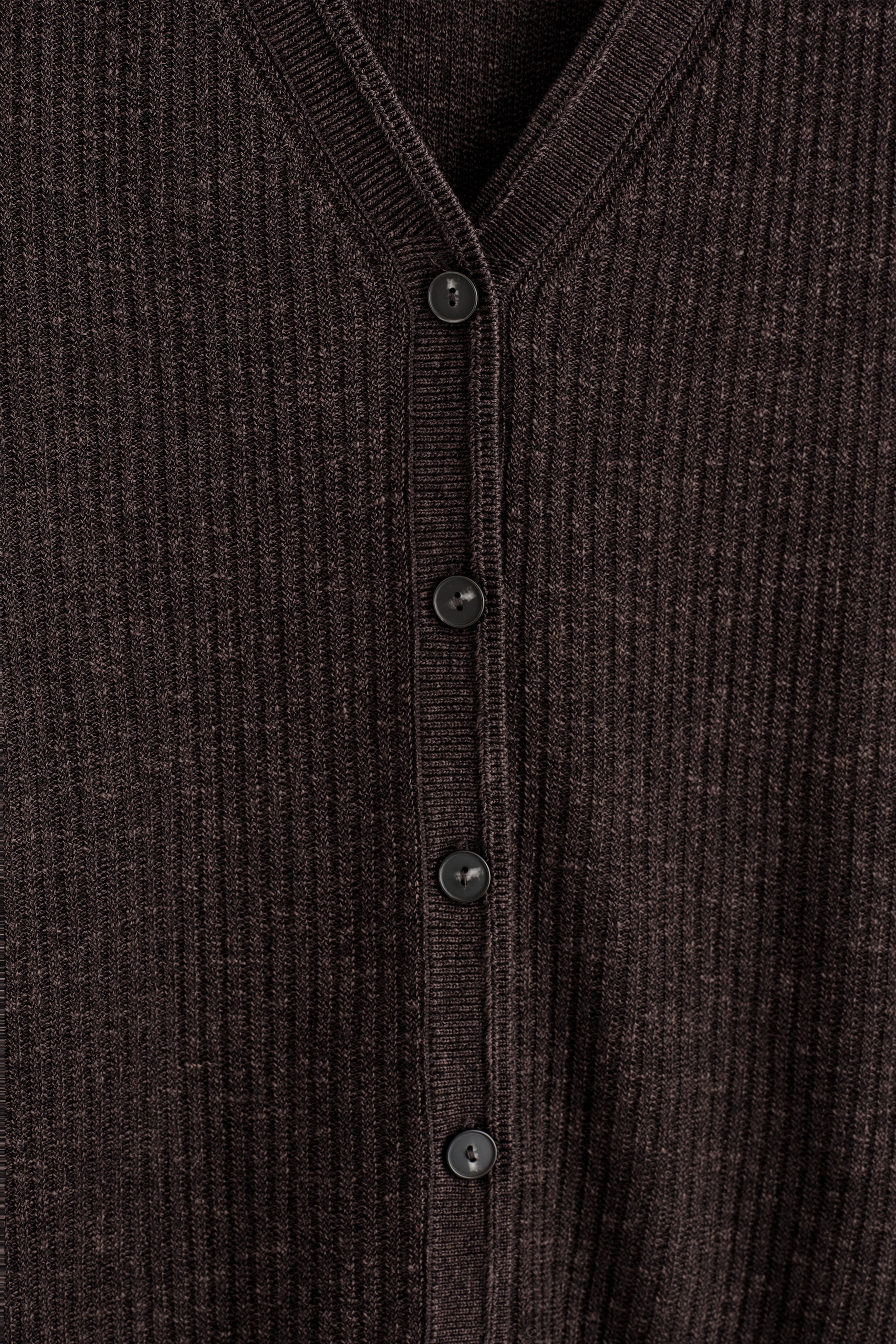Ingrandisci l'immagine: Un gilet a maglia a coste di tonalità scura viene presentato dalla parte anteriore. Presenta uno scollo a V e un'abbottonatura centrale con diversi bottoni scuri e circolari.