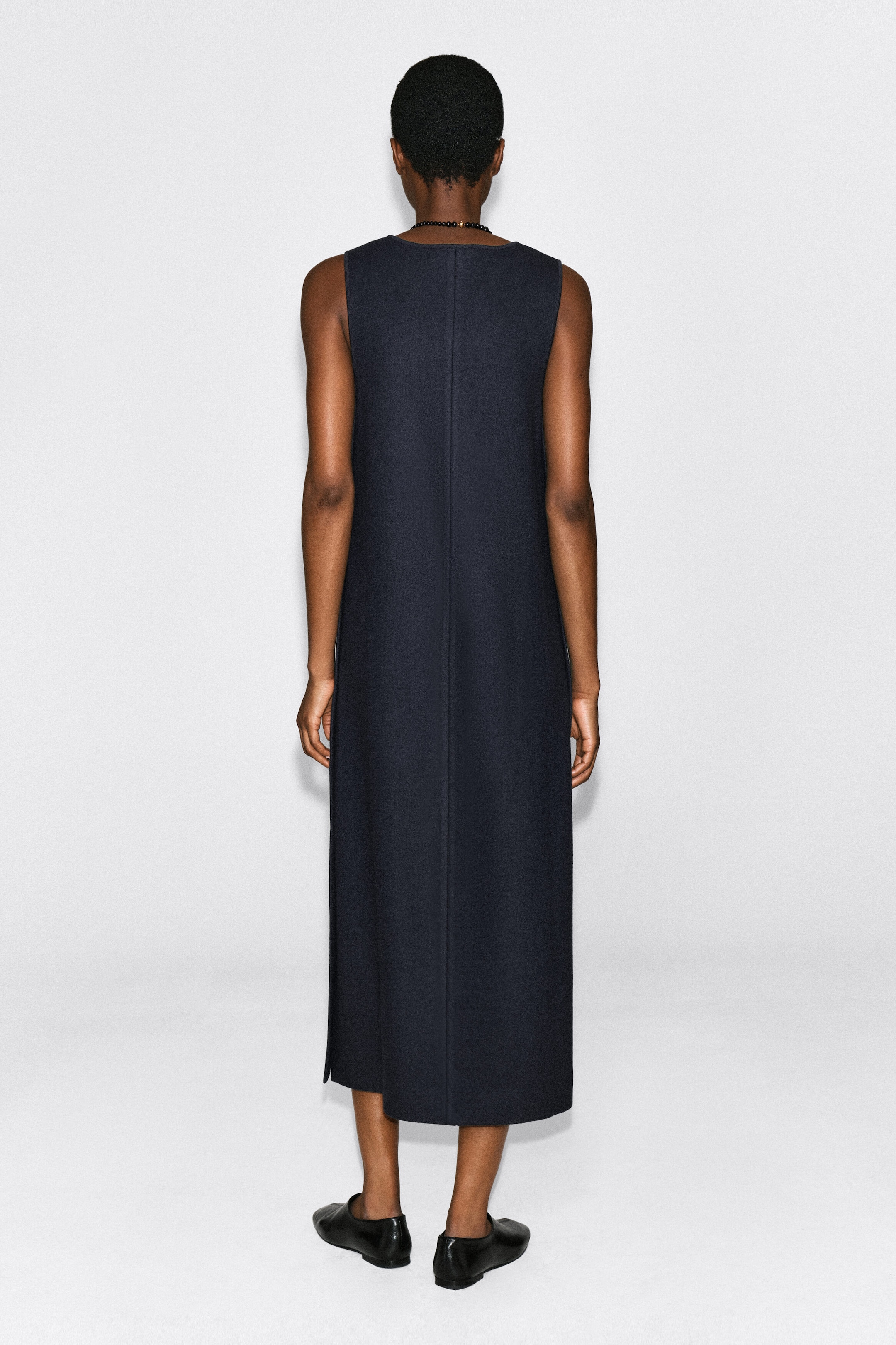 A dark navy boiled wool-blend midi dress is being worn by the model, shown from the back. The sleeveless, straight-cut dress features a clean center back seam and is styled with black leather flat shoes. A dark navy boiled wool-blend midi dress is being worn by the model, shown from the back. The sleeveless, straight-cut dress features a clean center back seam and is styled with black leather flat shoes.