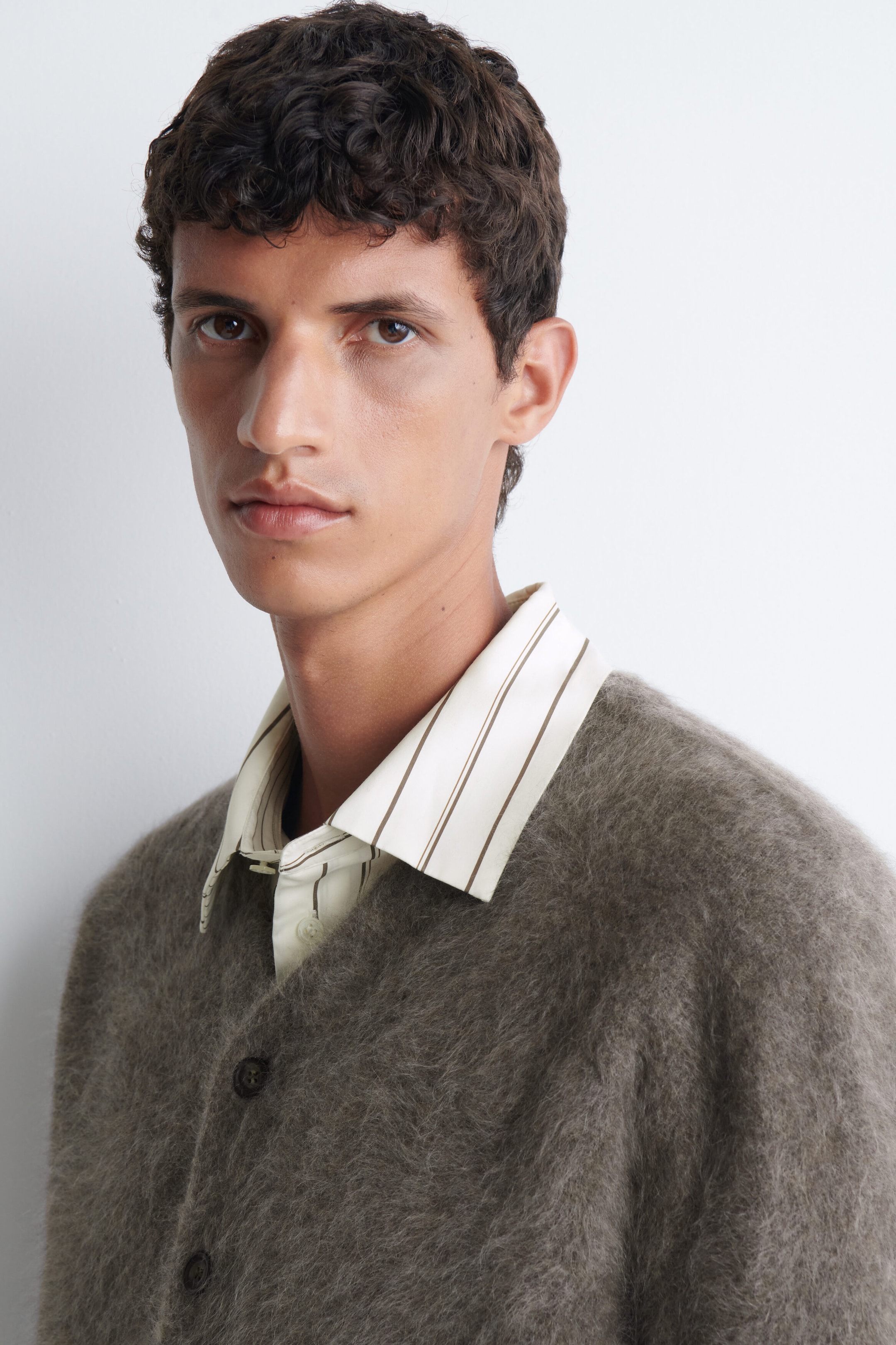 Un cardigan à col en V en cachemire brossé, dans un tissu taupe texturé et duveteux, est porté par le mannequin. Le cardigan est superposé à une chemise à col couleur crème, rehaussée de fines rayures verticales brunes. Un cardigan à col en V en cachemire brossé, dans un tissu taupe texturé et duveteux, est porté par le mannequin. Le cardigan est superposé à une chemise à col couleur crème, rehaussée de fines rayures verticales brunes.