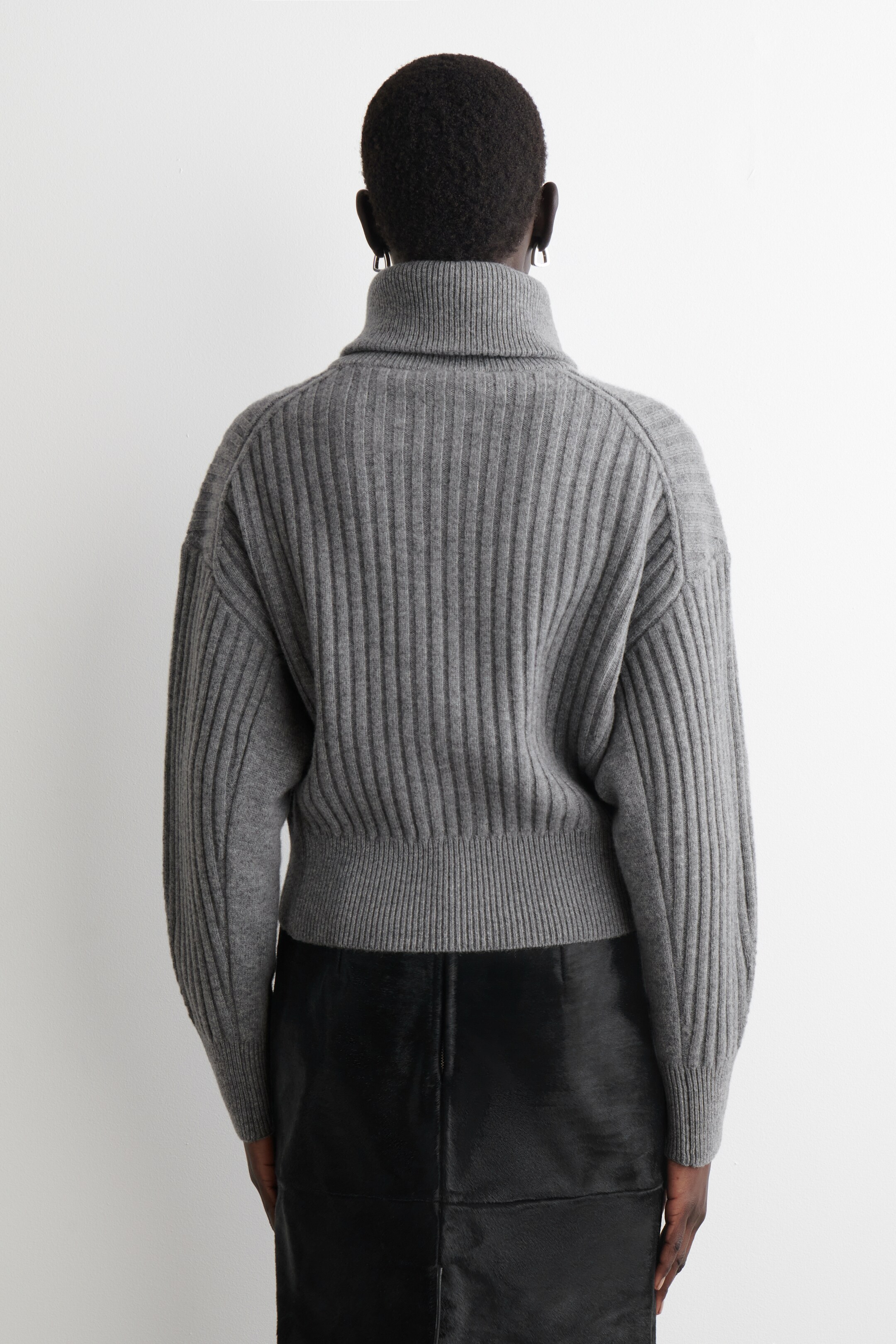 Größeres Bild ansehen: Ein grauer gerippter Rollkragenpullover wird von hinten präsentiert, mit auffälligen vertikalen Rippungen über den gesamten Körper und langen Ärmeln, einem hohen Rollkragen und einem gerippten Saum, der an der Taille endet.