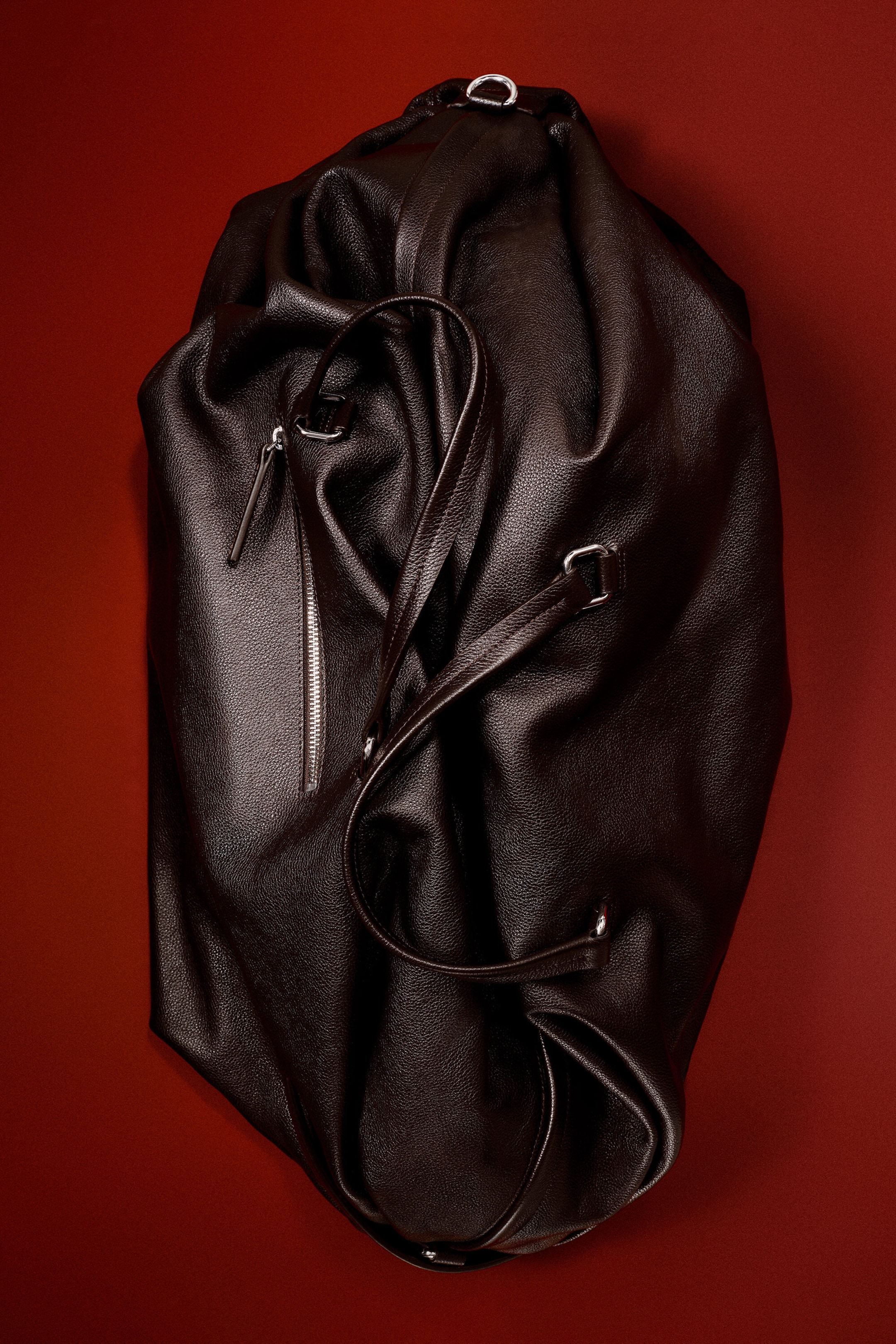 The Departure Duffle Bag in dark brown pebbled leather rests in a relaxed, unstructured silhouette, showcasing its soft drape and textured finish. It features dual leather top handles, a vertical silver-toned zippered pocket on the front, and metallic D-ring hardware.