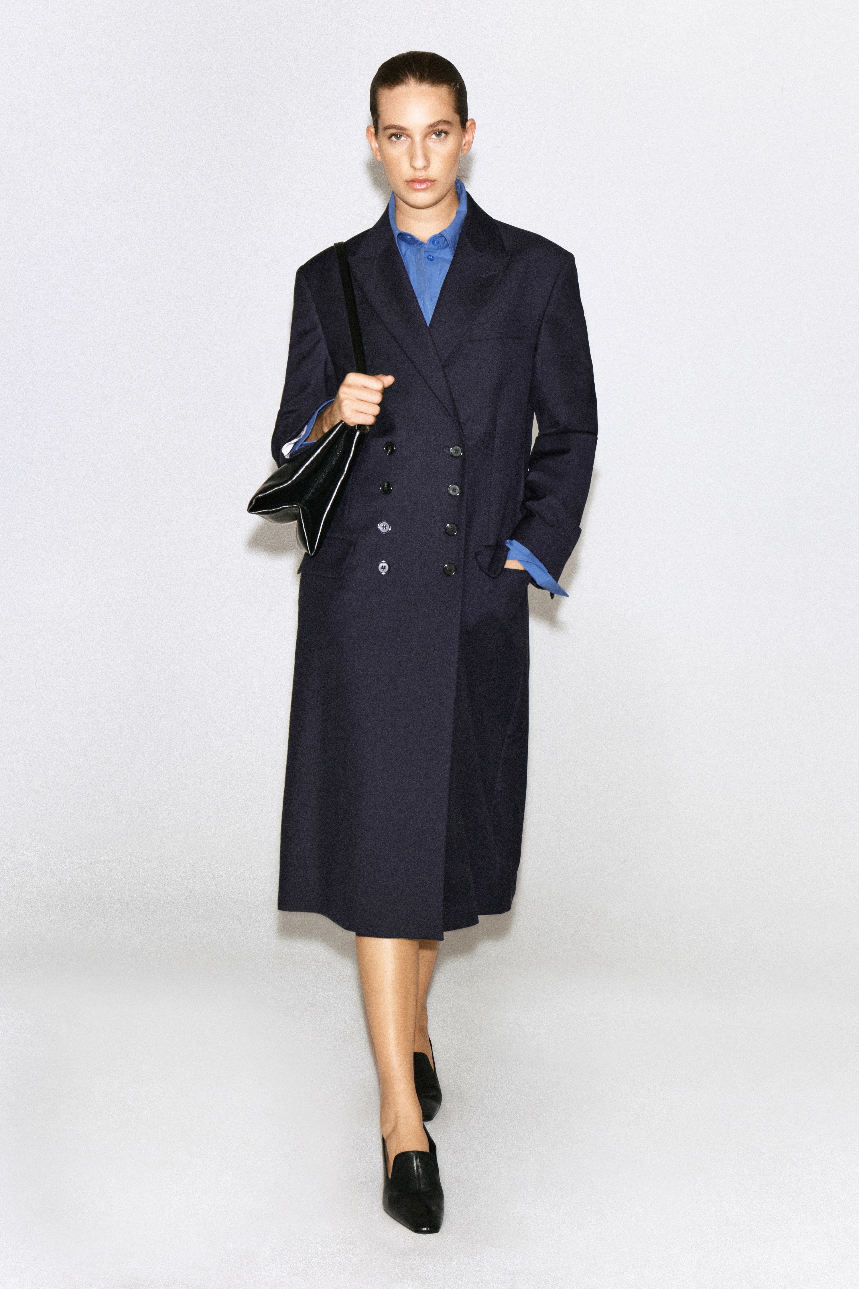 Um blazer comprido de trespasse duplo em azul-marinho escuro estÃ¡ pendurado de frente, destacando o seu design estruturado. O blazer apresenta lapelas de bico bem definidas, um bolso de ombreira no peito, dois bolsos laterais com aba e um fecho frontal com oito botÃµes.