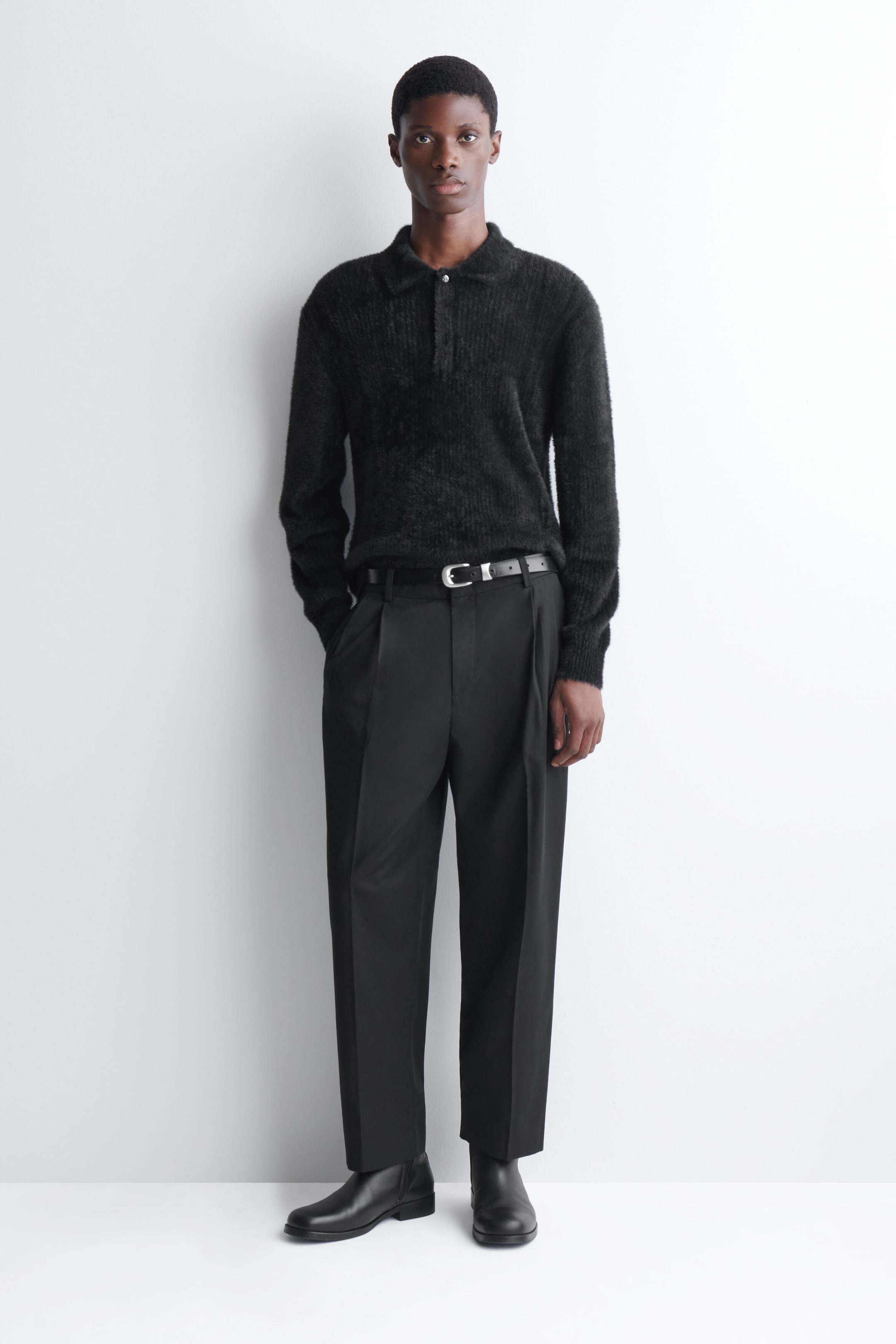The model is wearing a black knitted polo shirt with a textured, brushed finish, styled tucked into black tailored trousers with a belt. The long-sleeved shirt features a classic collar and ribbed cuffs. The model is wearing a black knitted polo shirt with a textured, brushed finish, styled tucked into black tailored trousers with a belt. The long-sleeved shirt features a classic collar and ribbed cuffs.