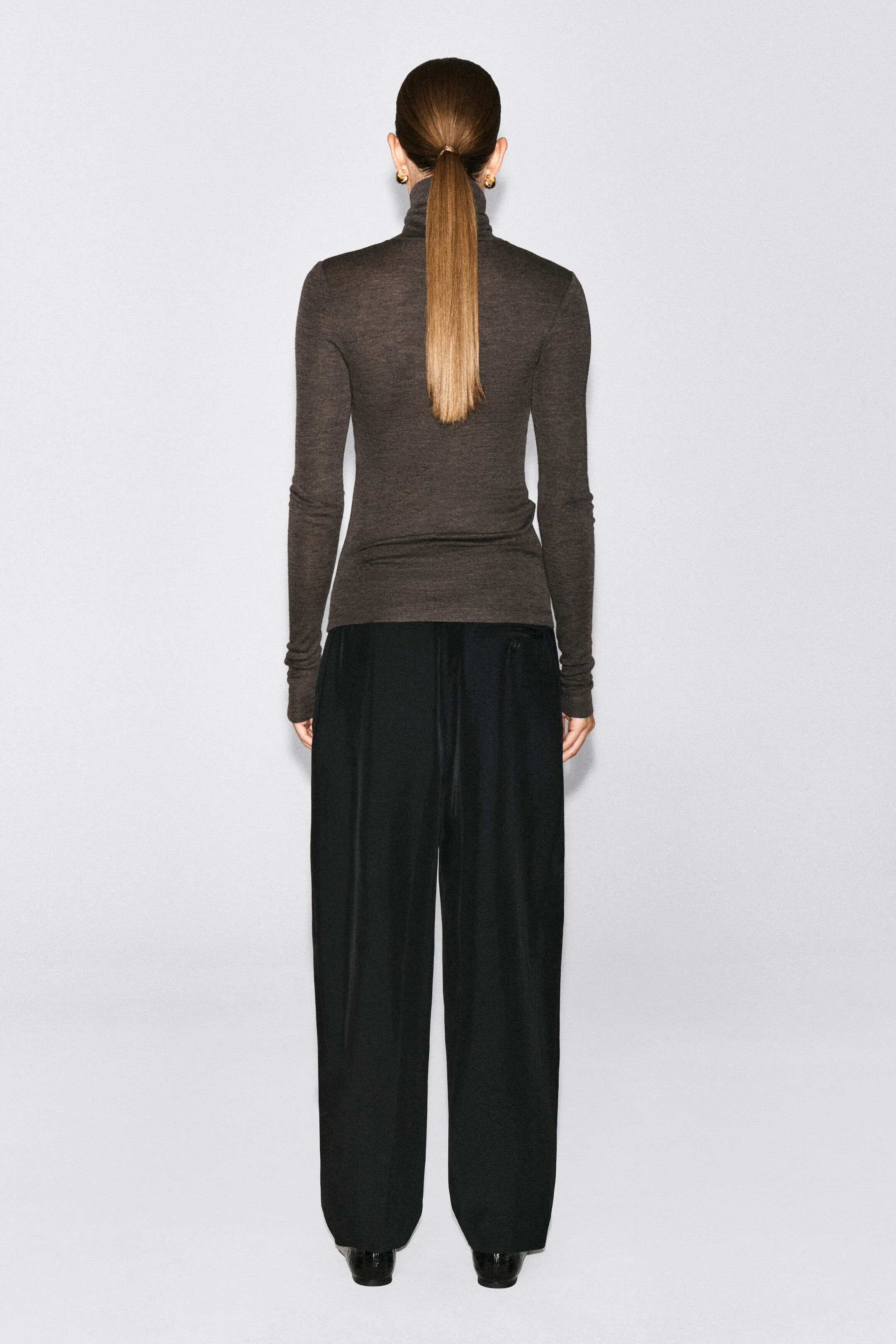 A dark brown merino wool high-neck top is being worn by the model, shown from the back. The slim-fitting, long-sleeved top is paired with wide-leg black trousers. A dark brown merino wool high-neck top is being worn by the model, shown from the back. The slim-fitting, long-sleeved top is paired with wide-leg black trousers.