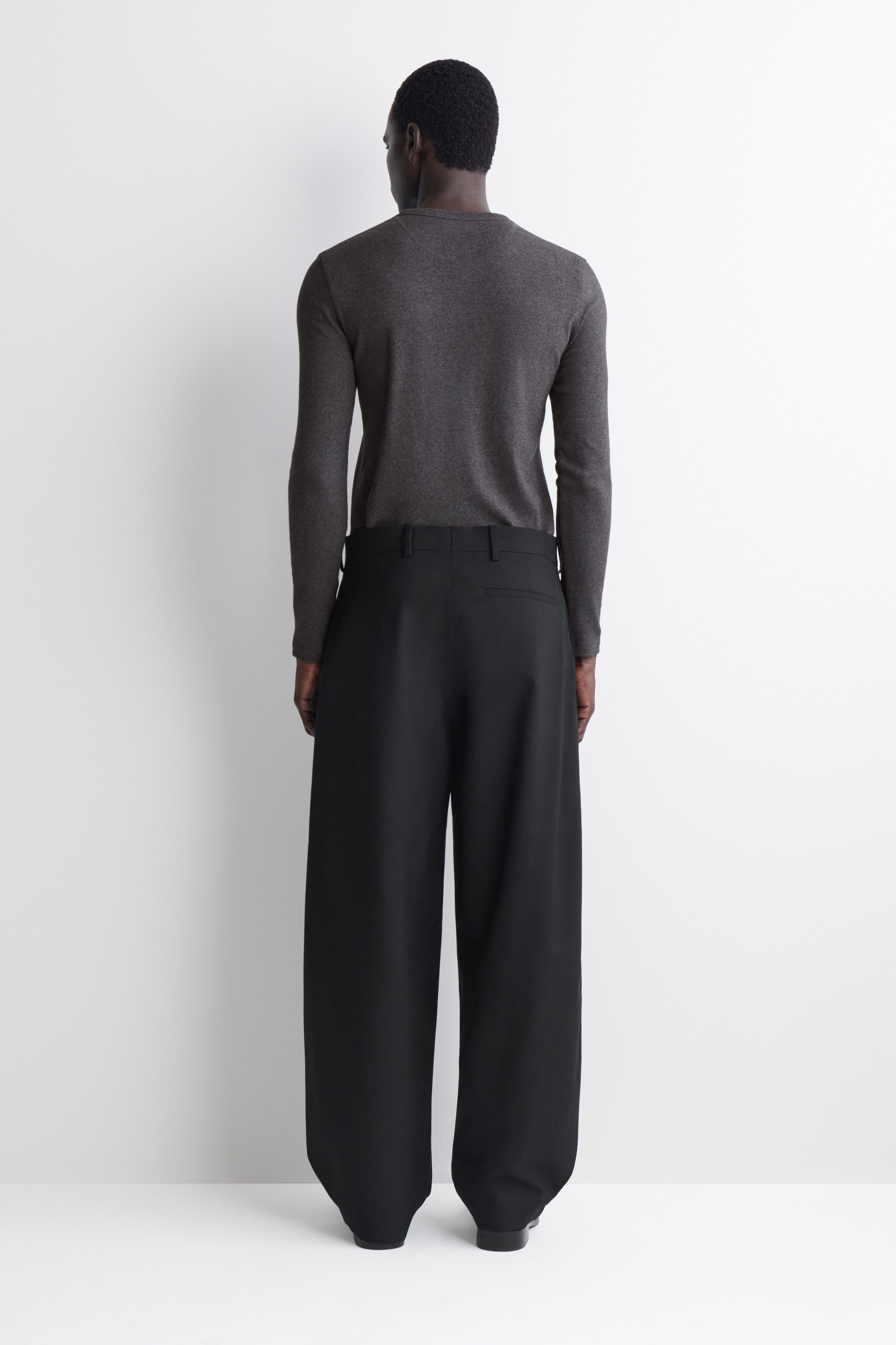 This slim, ribbed cotton long-sleeve Henley T-shirt is shown from the back, highlighting its close-fitting silhouette and dark gray ribbed fabric. The T-shirt is styled tucked into wide-leg black trousers to emphasize its slim, structured fit. This slim, ribbed cotton long-sleeve Henley T-shirt is shown from the back, highlighting its close-fitting silhouette and dark gray ribbed fabric. The T-shirt is styled tucked into wide-leg black trousers to emphasize its slim, structured fit.