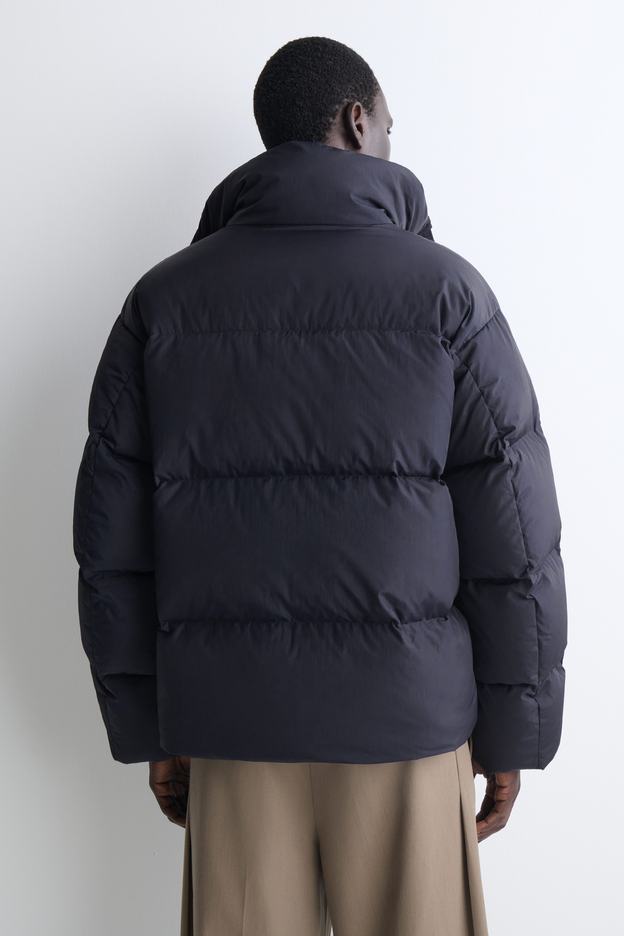 This dark gray funnel-neck nylon down puffer jacket is shown from the back, highlighting its voluminous, relaxed fit. It features horizontal quilting across the body and sleeves, along with a high, padded collar.
