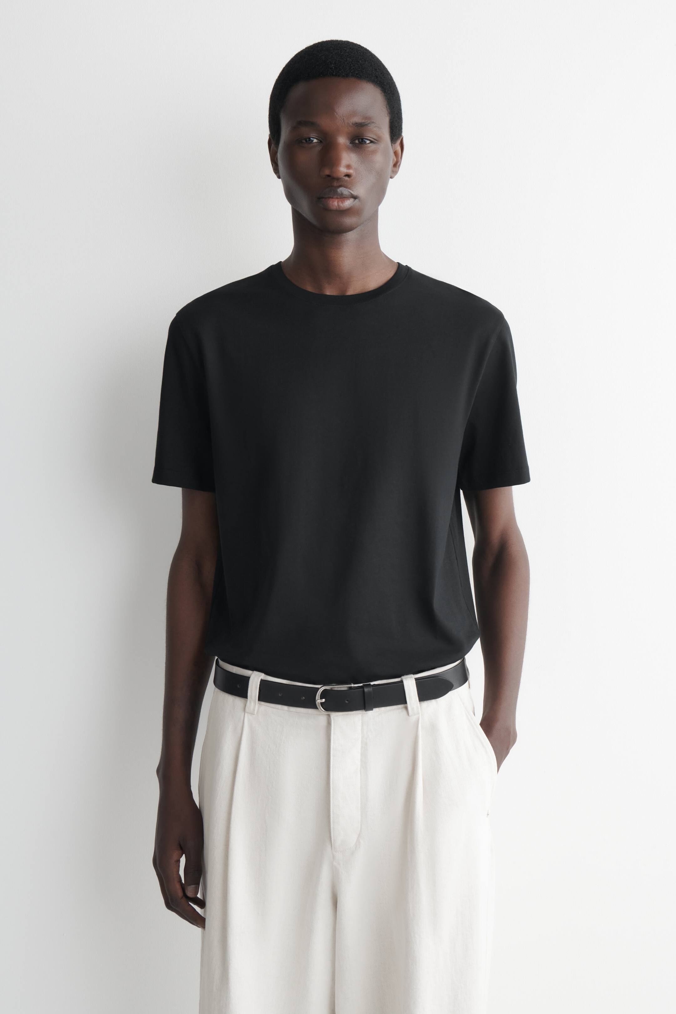 Visualizza immagine più grande: La modella indossa una t-shirt nera a maniche corte con scollo rotondo. È infilata in pantaloni a pieghe di colore chiaro e fissata con una cintura nera.