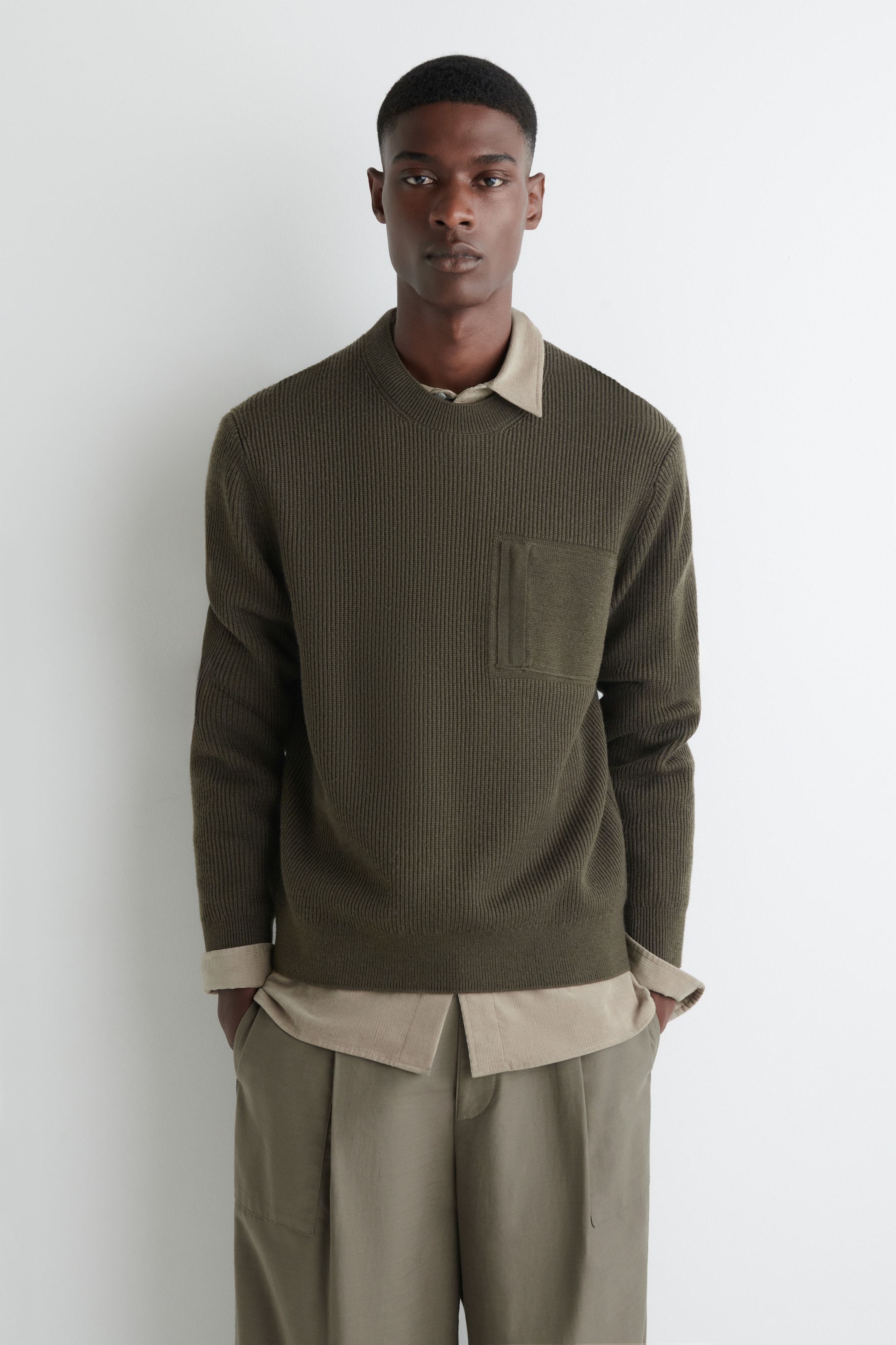 Agrandir l'image: Un pull en tricot côtelé vert olive est présenté de face, avec un col rond, une poche plaquée texturée sur la poitrine gauche et un ourlet côtelé. Une chemise à col de couleur plus claire est superposée en dessous, visible au niveau de l'encolure et des poignets.