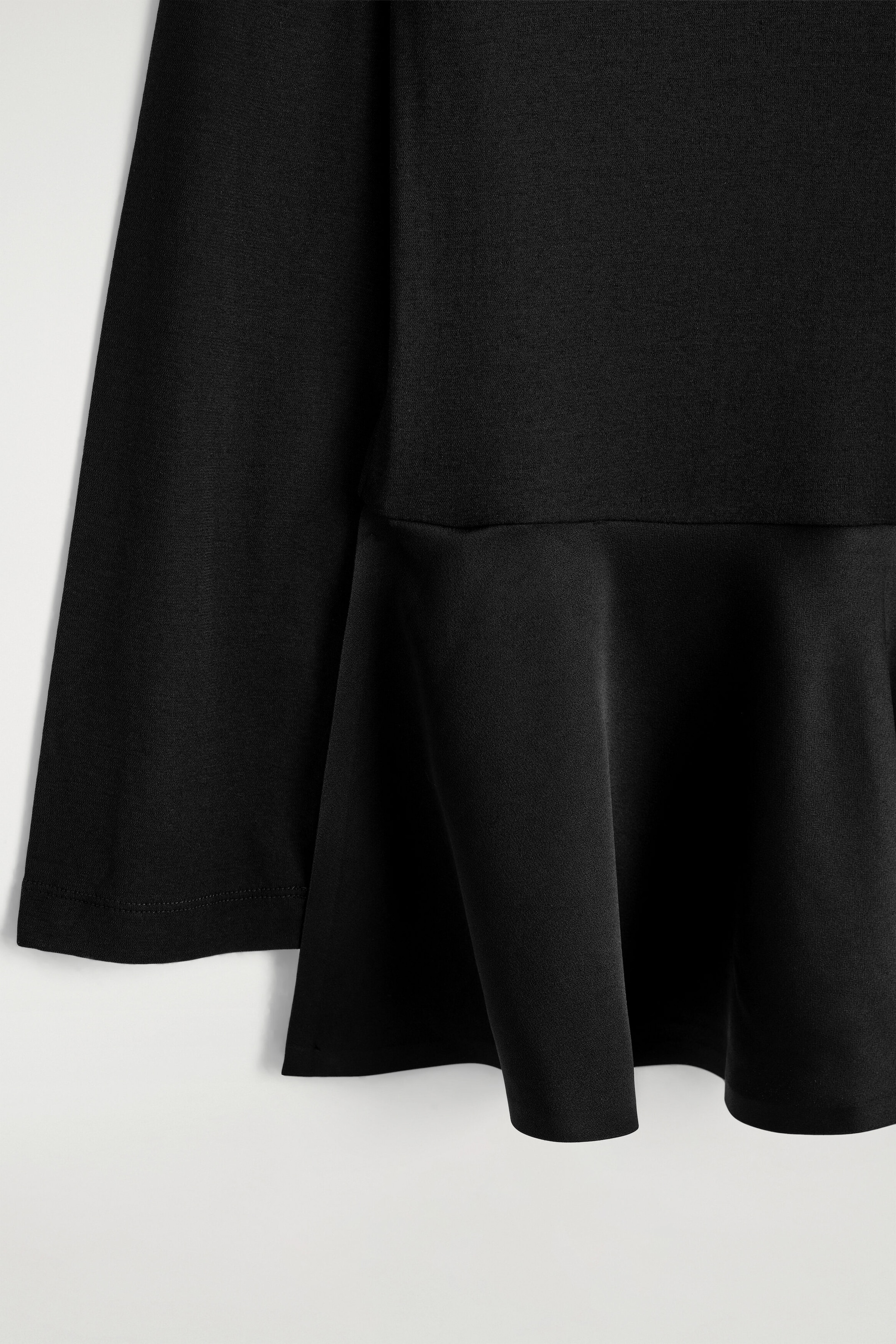 Le haut en jersey à basque satinée est présenté en vue rapprochée de face, mettant en valeur un corsage en jersey mat uni à un ourlet basque évasé en satin drapé. Une manche longue avec poignet cousu retombe sur le côté gauche, à côté des plis fluides de l’ourlet.