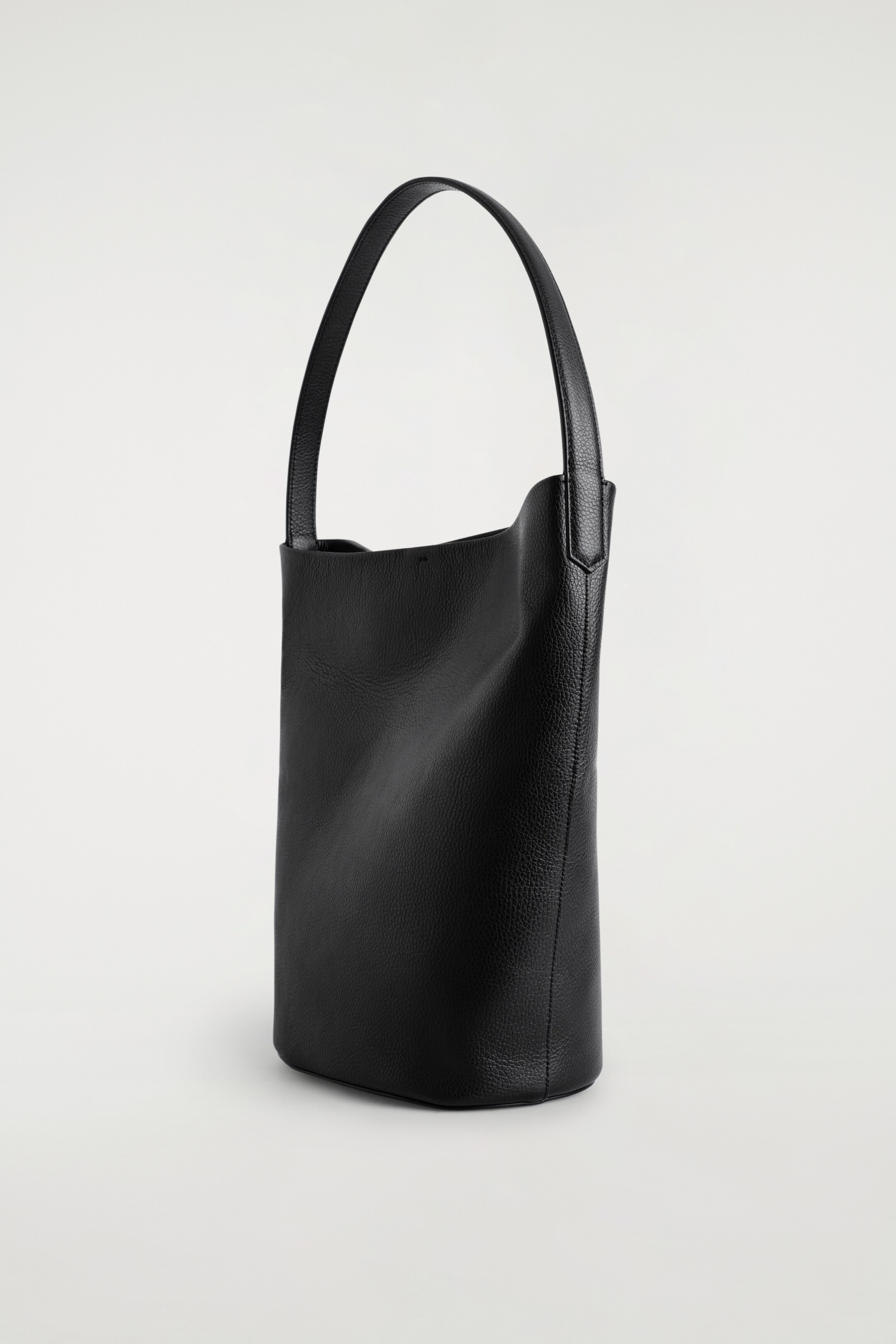 Ver imagem maior: Uma mala tote de couro preto e texturizado está em PÉ, apresentando uma silhueta suave e desleixada de balde que se dobra suavemente para dentro no topo. Uma única alça esguia está arqueando graciosamente das bordas superiores da mala.