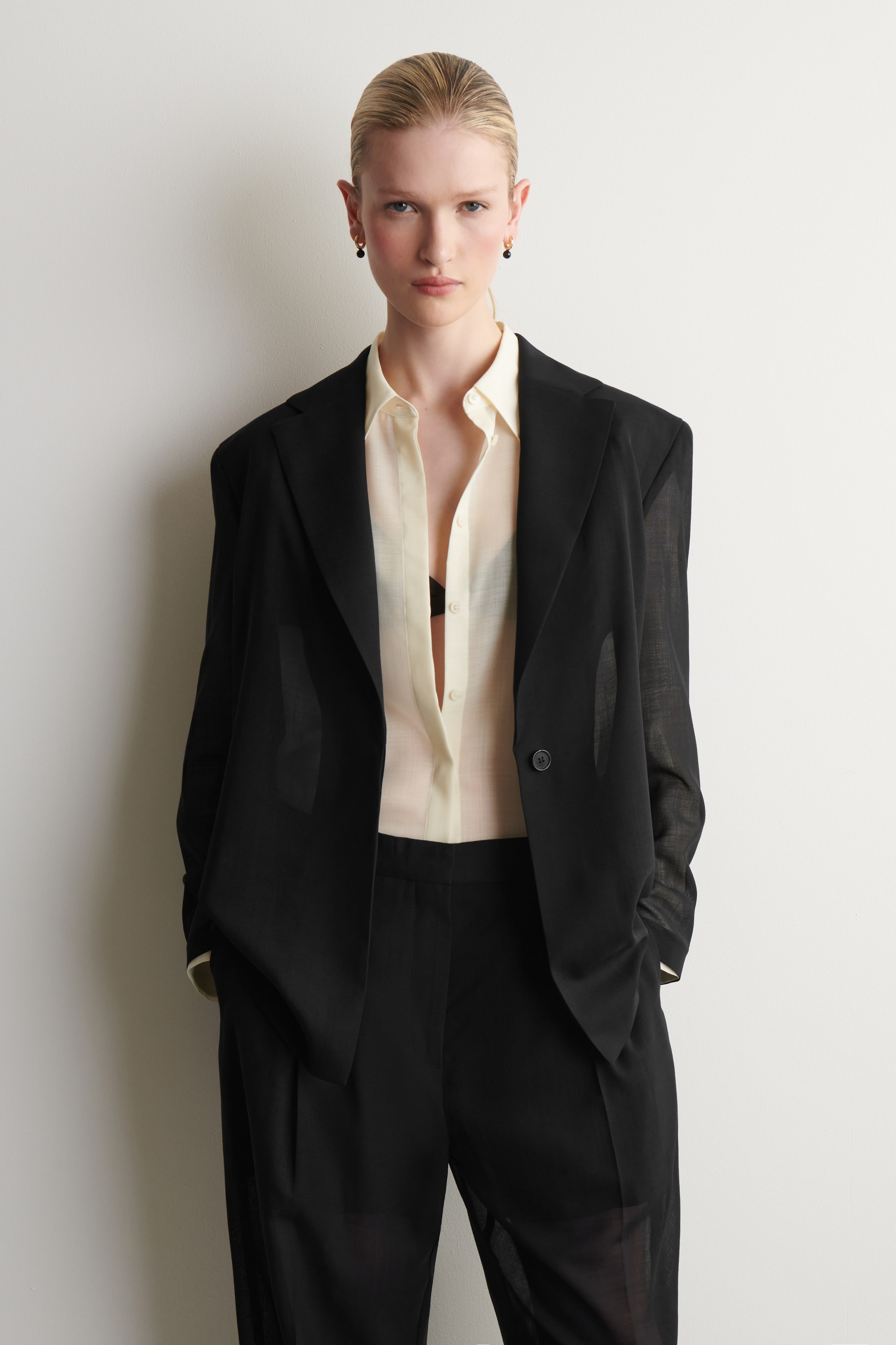 Visualizza l'immagine ingrandita: Un blazer nero e pantaloni coordinati sono indossati, rivolti in avanti. Il blazer presenta revers a lancia, una chiusura a un bottone e un tessuto leggero che è leggermente trasparente. È aperto sopra una camicia con colletto di colore chiaro, e i pantaloni abbinati presentano una vestibilità rilassata e a lunghezza intera.