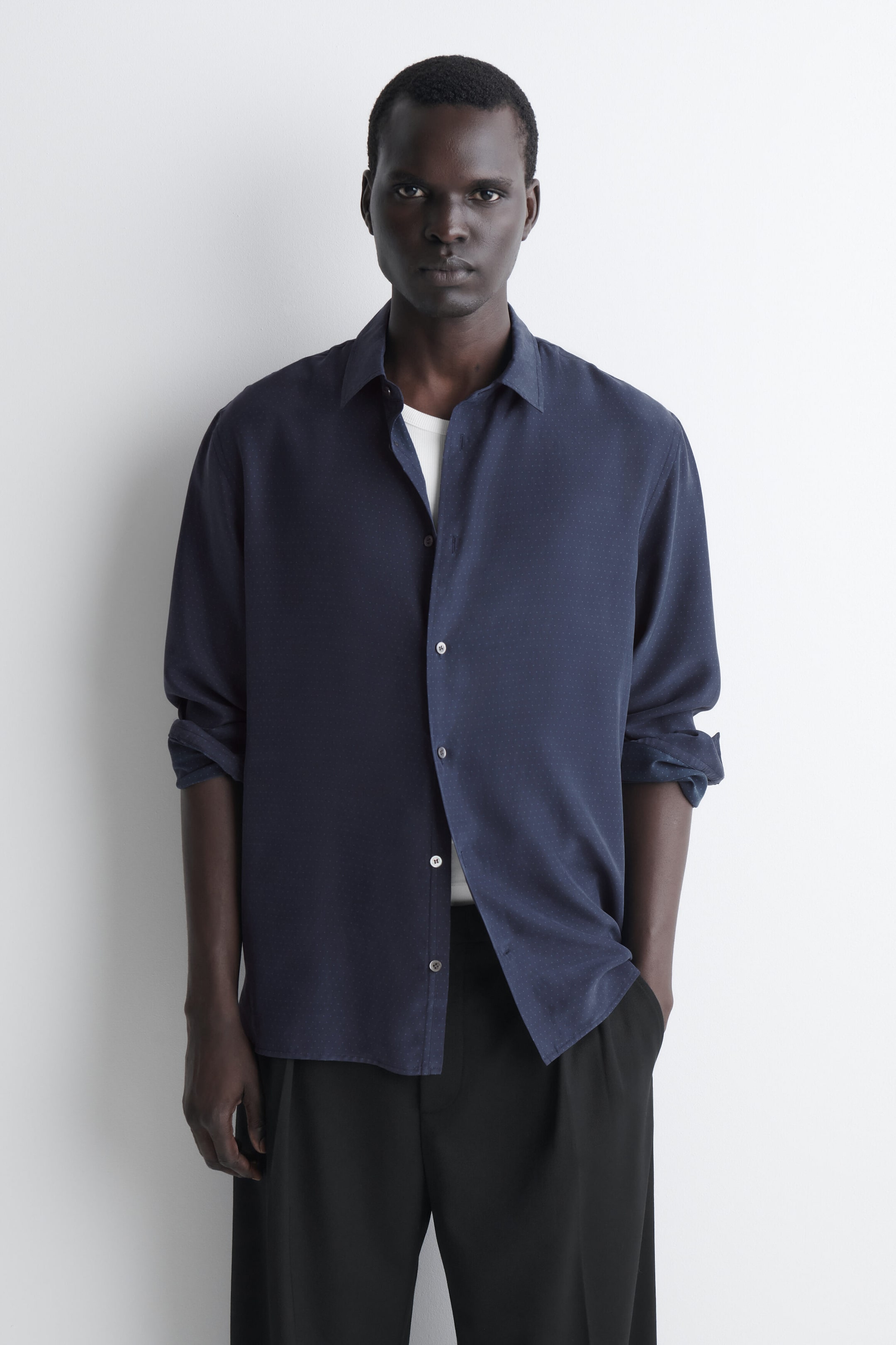 The navy blue sandwashed silk shirt is shown from the front in a relaxed fit, featuring a standard collar, button-down front, and subtle micro-dot texture. It is layered over a white T-shirt, with the long sleeves rolled up to the elbows. The navy blue sandwashed silk shirt is shown from the front in a relaxed fit, featuring a standard collar, button-down front, and subtle micro-dot texture. It is layered over a white T-shirt, with the long sleeves rolled up to the elbows.
