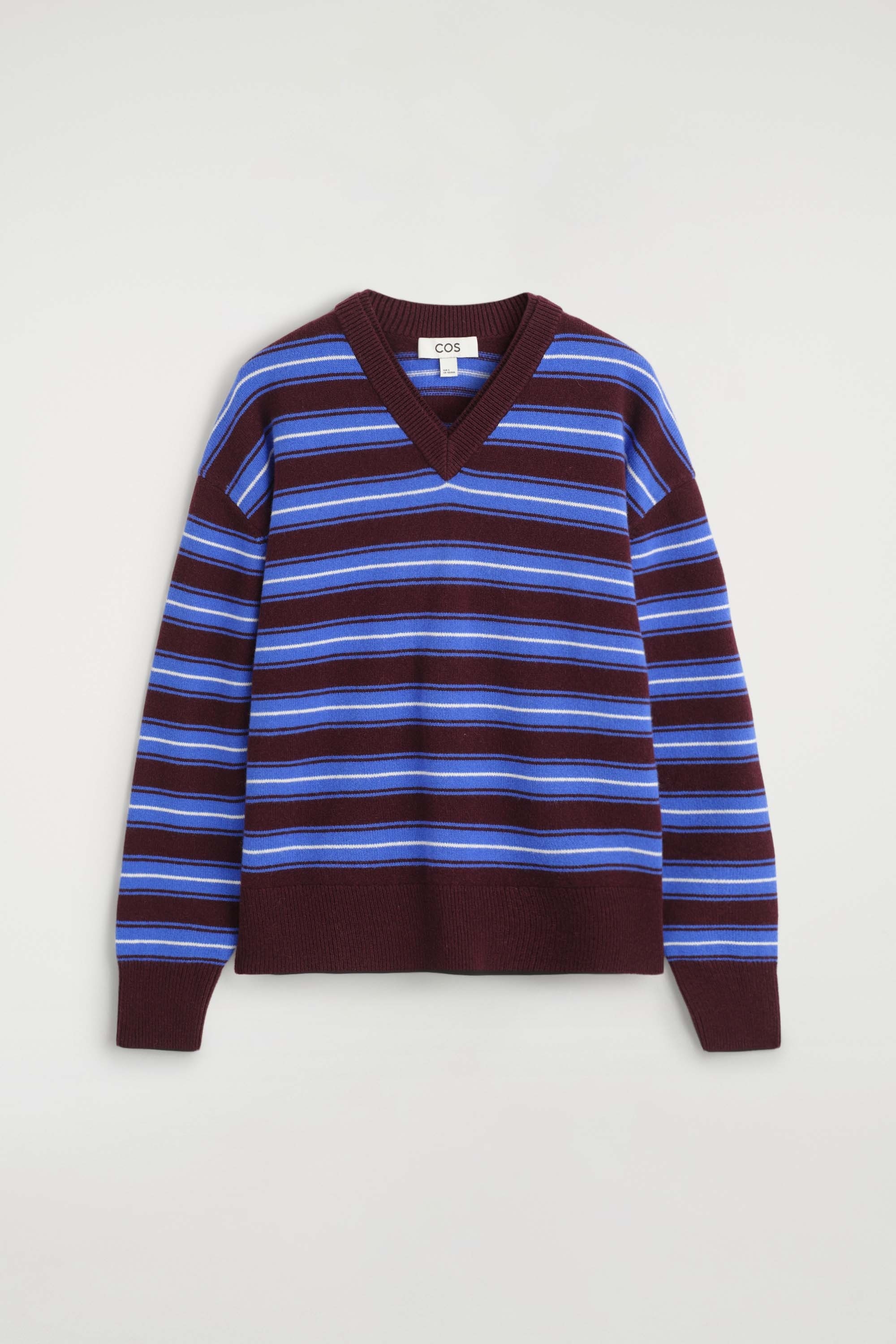 The merino wool layered V-neck jumper, featuring horizontal stripes in blue, white and burgundy, is worn by the model. The jumper is styled with a black midi wrap skirt, black sunglasses and black slip-on shoes, highlighting its relaxed fit, ribbed burgundy cuffs and double-layered V-neckline.