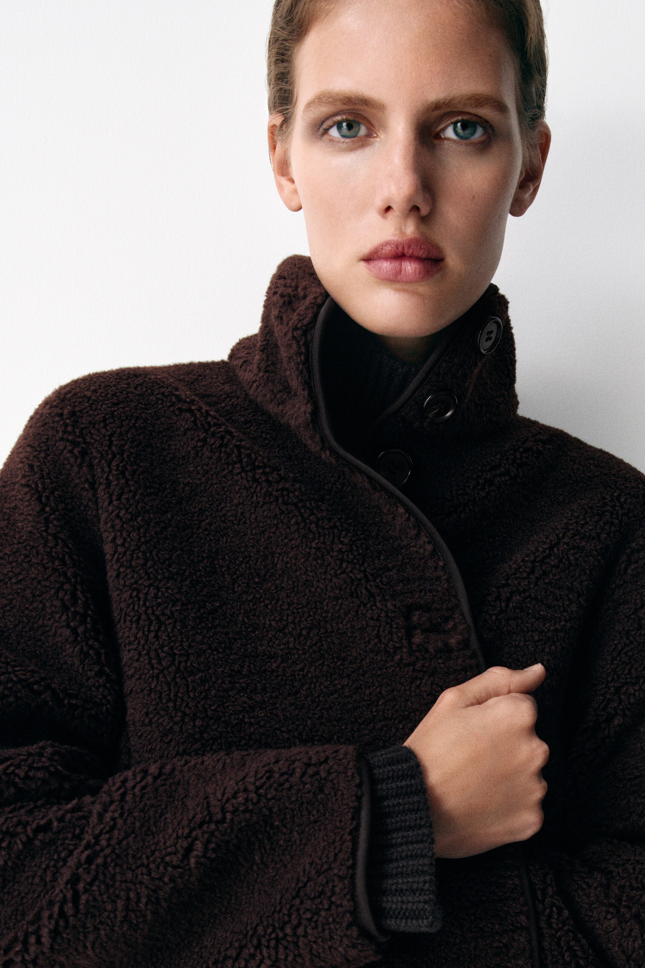 Größeres Bild anzeigen: Eine dunkelbraune Jacke mit Trichterkragen zeigt ihre plüschige Teddy-Textur und verfügt über einen Verschluss mit schwarzen Knöpfen und tonaler Einfassung entlang der Kanten. Ein dunkles geripptes Bündchen ist sichtbar unter dem Ärmel der Jacke geschichtet.