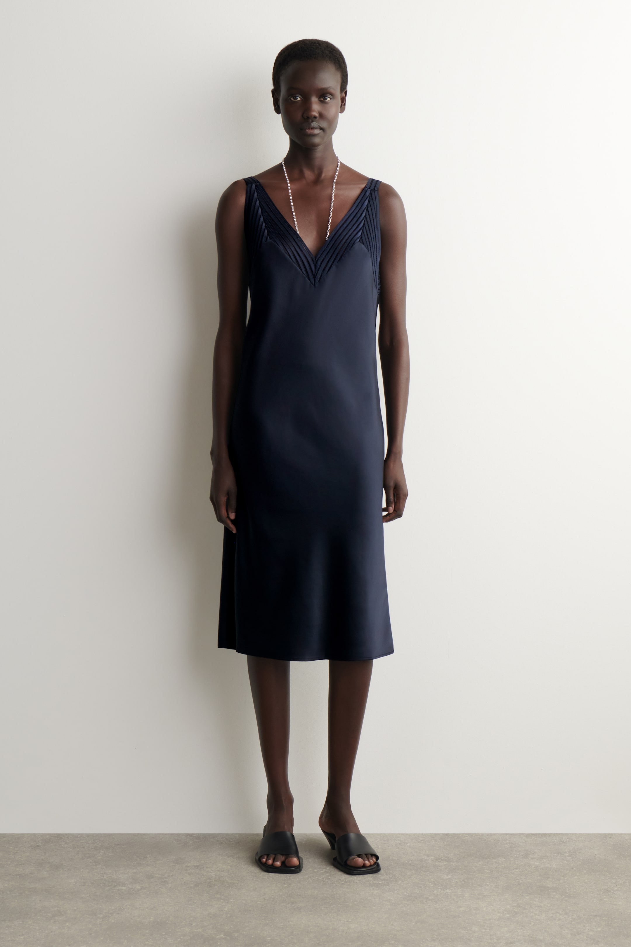 Agrandir l'image: Une robe midi bleu foncé dotée d'un décolleté en V avec des détails plissés en forme de V sur le corsage, de fines bretelles et une silhouette fluide s'étendant juste en dessous du genou.