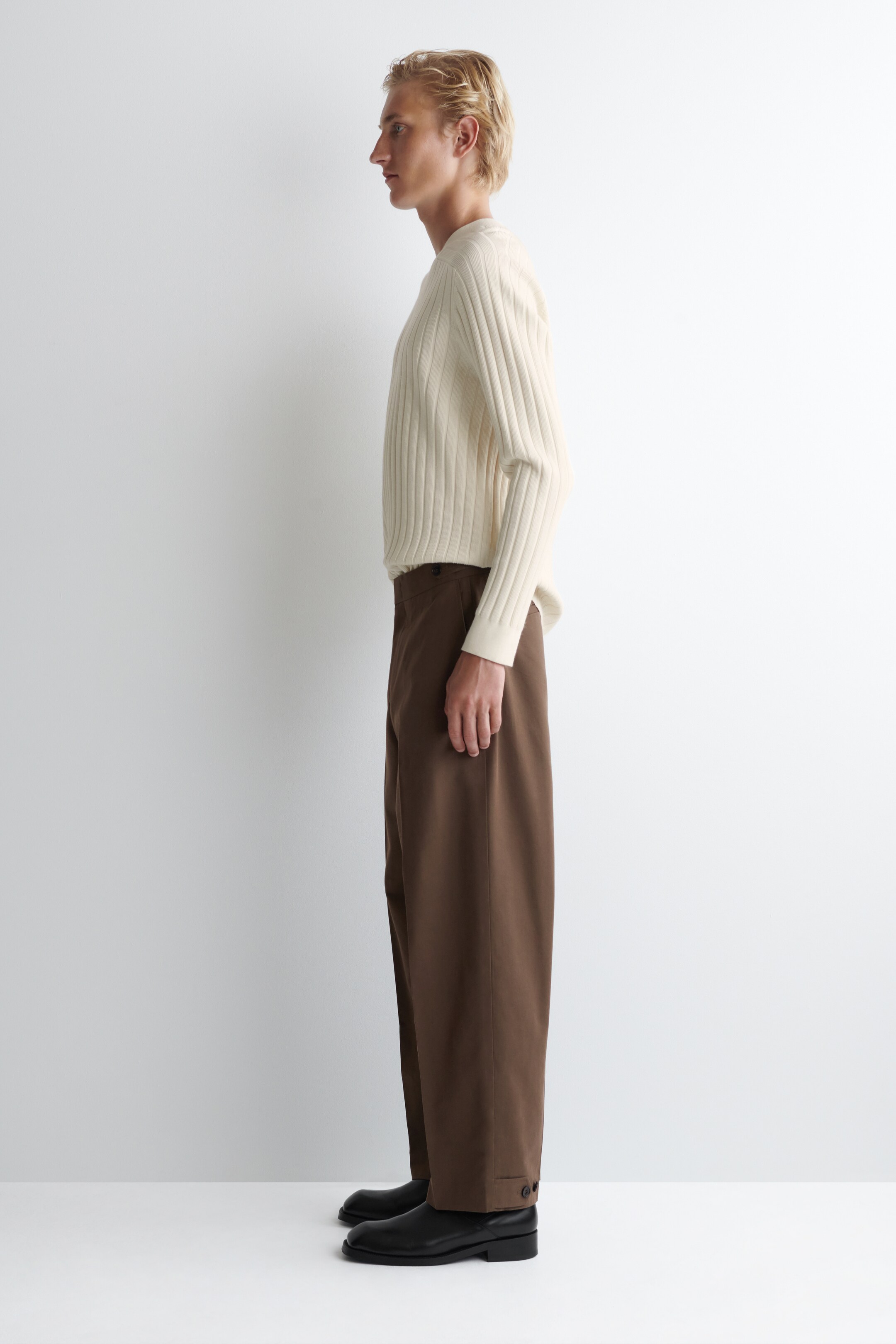 Agrandir l'image: Un pull crème à côtes verticales avec un col rond et des poignets côtelés est porté, visible de côté. Il est associé à un pantalon marron à jambes larges avec un détail de patte boutonnée près de l'ourlet, et des chaussures noires complètent l'ensemble.