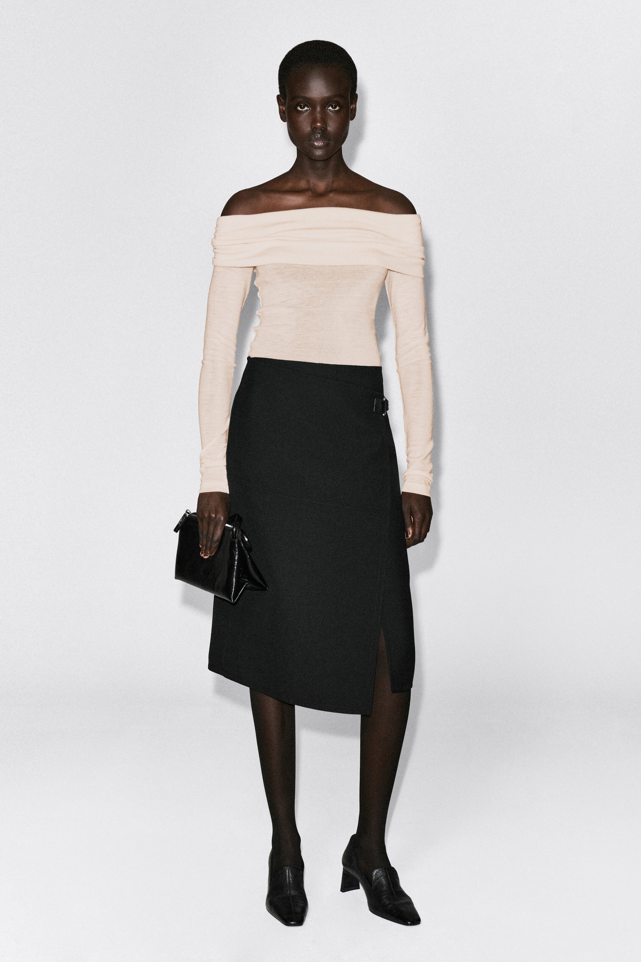 An off-the-shoulder merino wool top in a light beige color is shown from the front, featuring a wide, folded-over draped neckline and long sleeves. The semi-sheer, fine-knit top fits closely to the body and is styled tucked into a black wrap midi skirt. An off-the-shoulder merino wool top in a light beige color is shown from the front, featuring a wide, folded-over draped neckline and long sleeves. The semi-sheer, fine-knit top fits closely to the body and is styled tucked into a black wrap midi skirt.