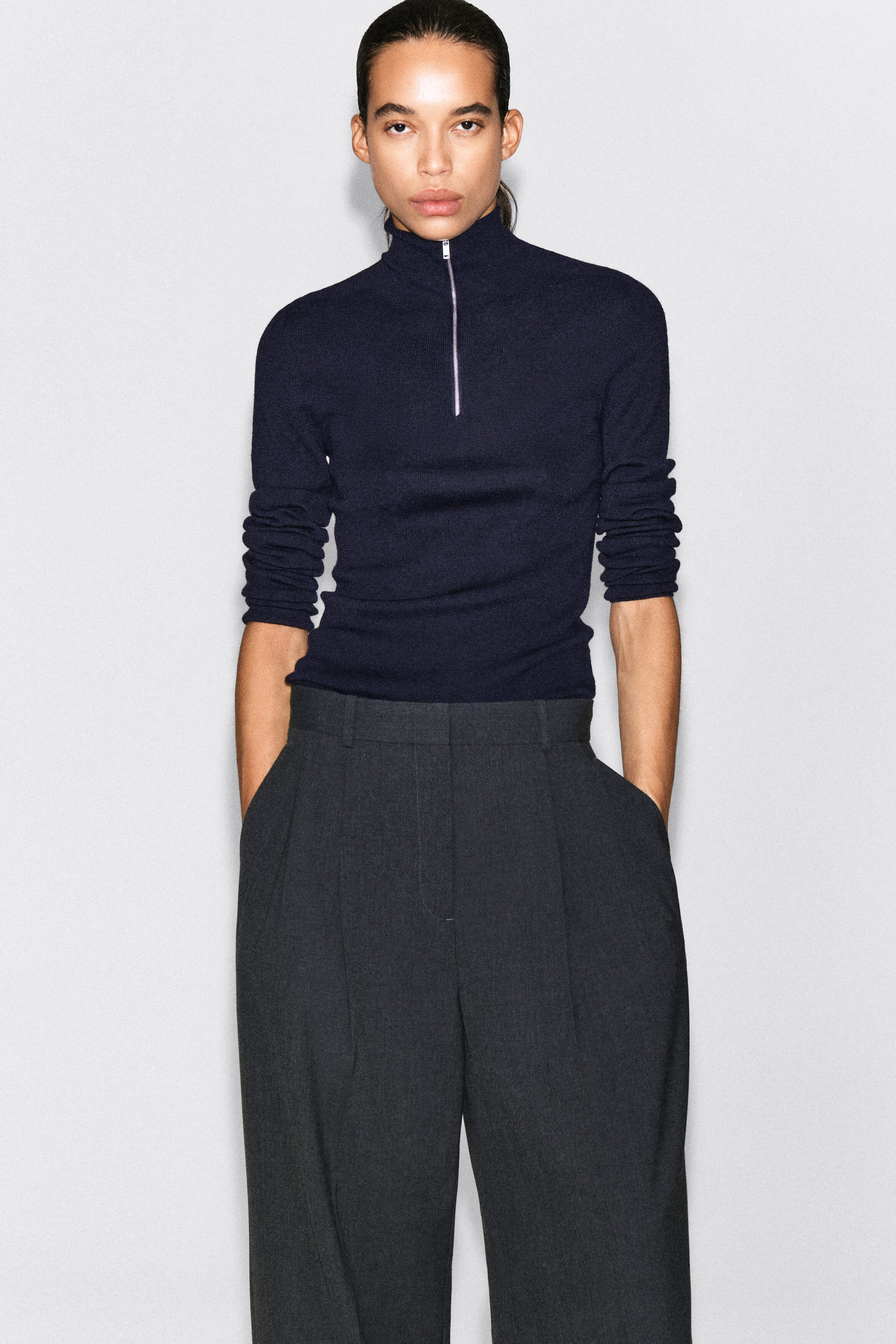 A dark navy blue slim-fit mock-neck top features a silver-tone half-zip closure and long sleeves pushed up to the forearms. The top is styled tucked into high-waisted, pleated dark grey trousers. A dark navy blue slim-fit mock-neck top features a silver-tone half-zip closure and long sleeves pushed up to the forearms. The top is styled tucked into high-waisted, pleated dark grey trousers.