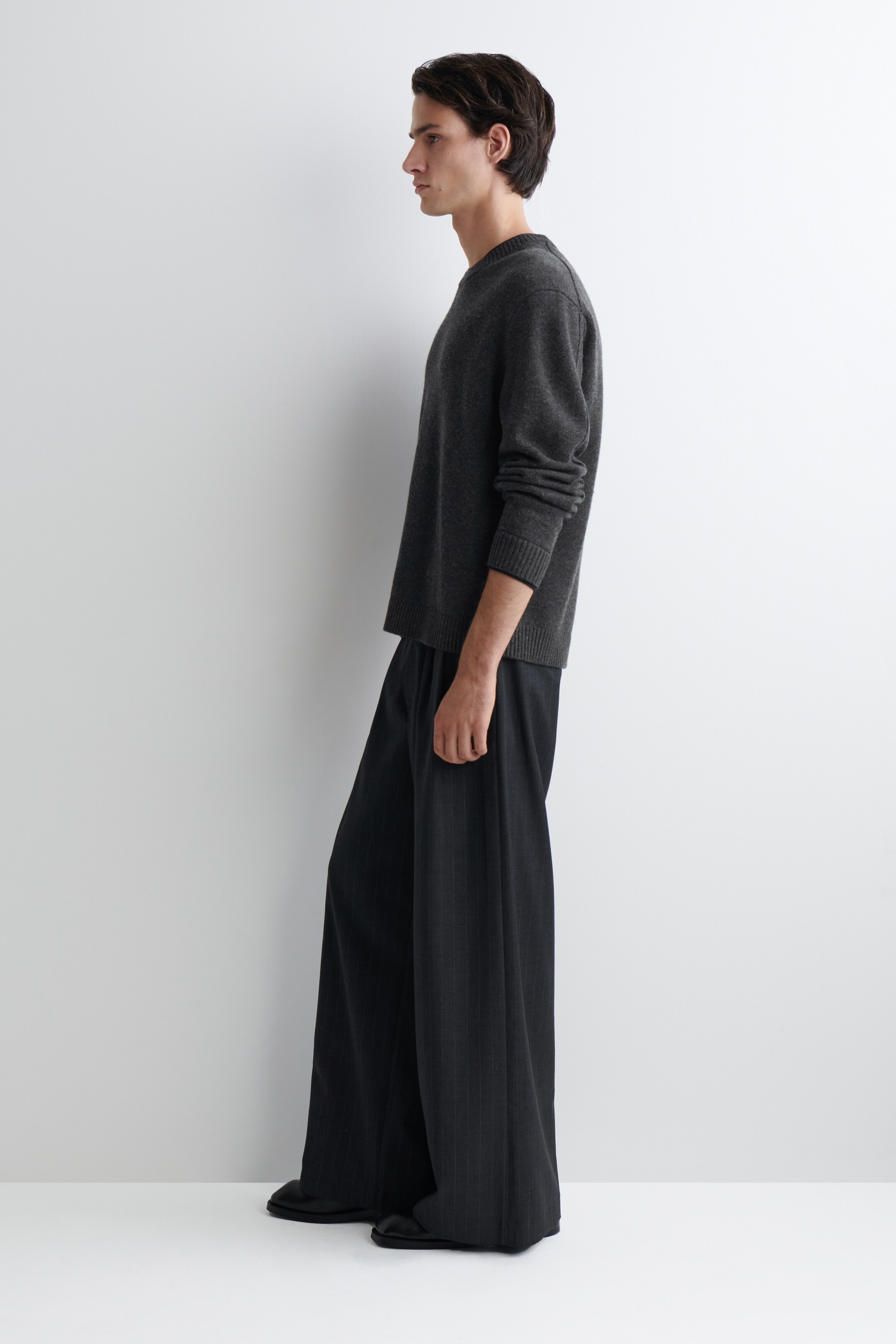 View larger image: A charcoal gray crew neck sweater with ribbed cuffs and hem is being worn, showcasing its relaxed fit from a side profile. It is being paired with dark gray wide-leg trousers, also seen from the side, which are displaying a flowing silhouette and subtle vertical pinstripes.