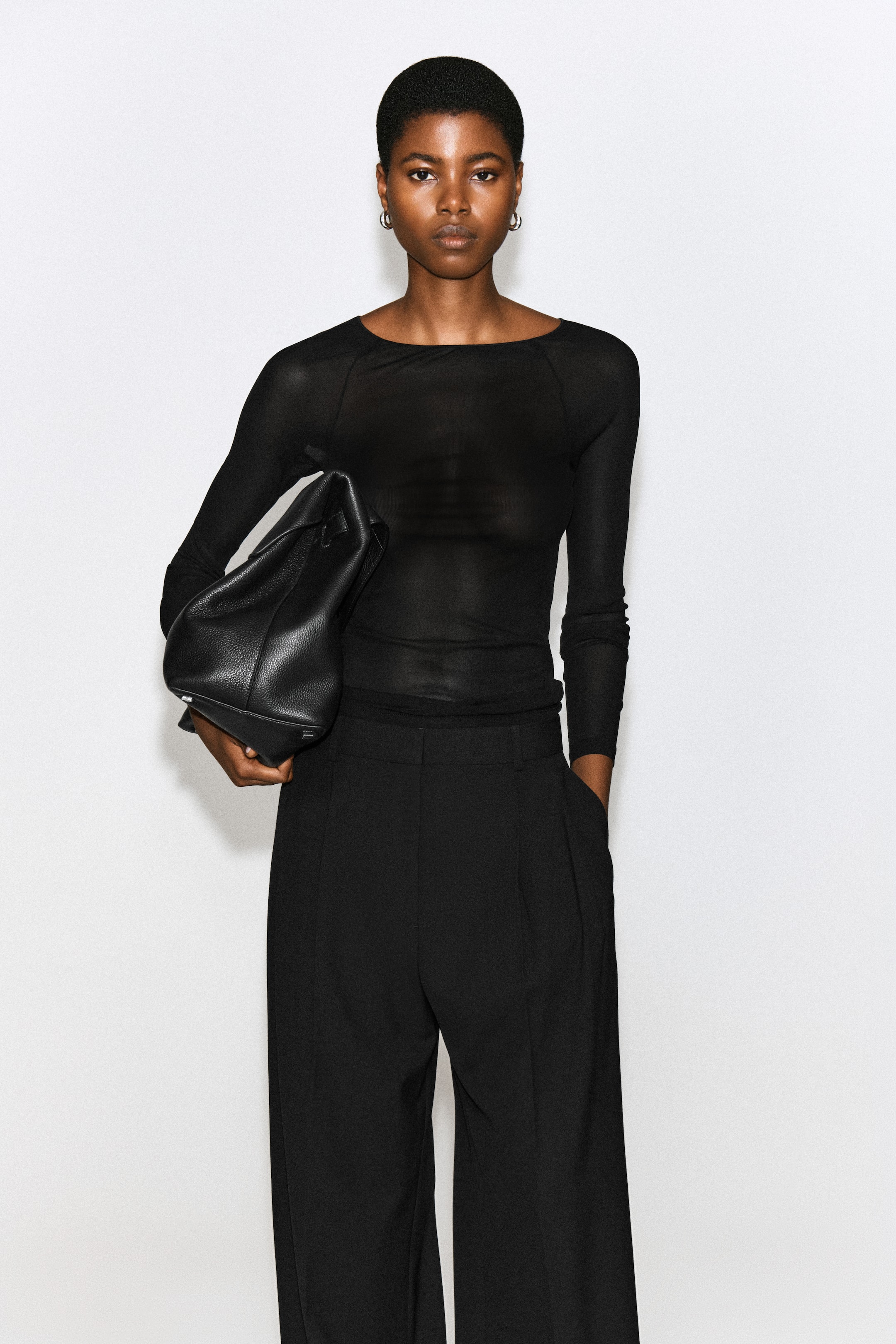 Ver imagen más grande: La modelo lleva un top de seda negro entallado con canalé, de textura semitransparente, mangas largas y cuello barco. El top está metido por dentro de un pantalón negro de corte sastre, en combinación con un bolso grande de piel negra que lleva bajo el brazo.